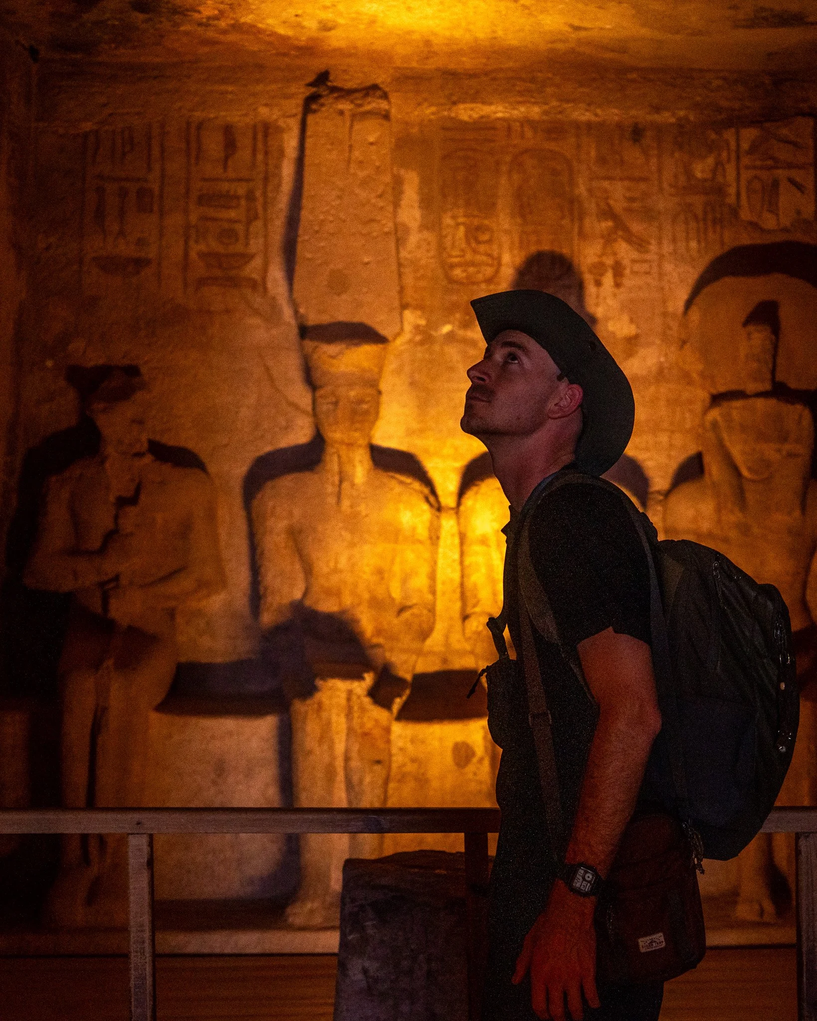 A man in a black shirt, backpack, and cowboy hat looking up at ancient Egyptian hieroglyphs and statues illuminated by warm lighting in a museum.