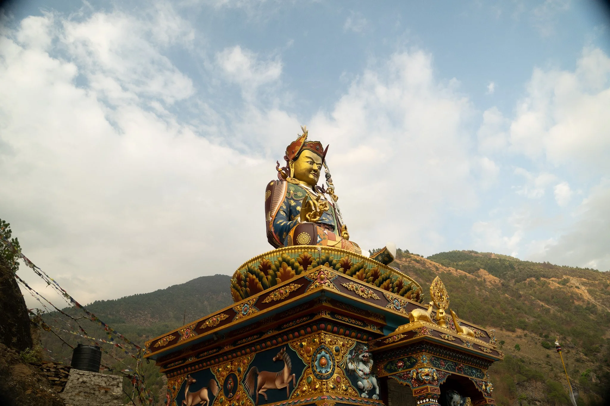 A large, colorful statue of a deity or spiritual figure seated on a decorated platform, with mountains and a cloudy sky in the background.