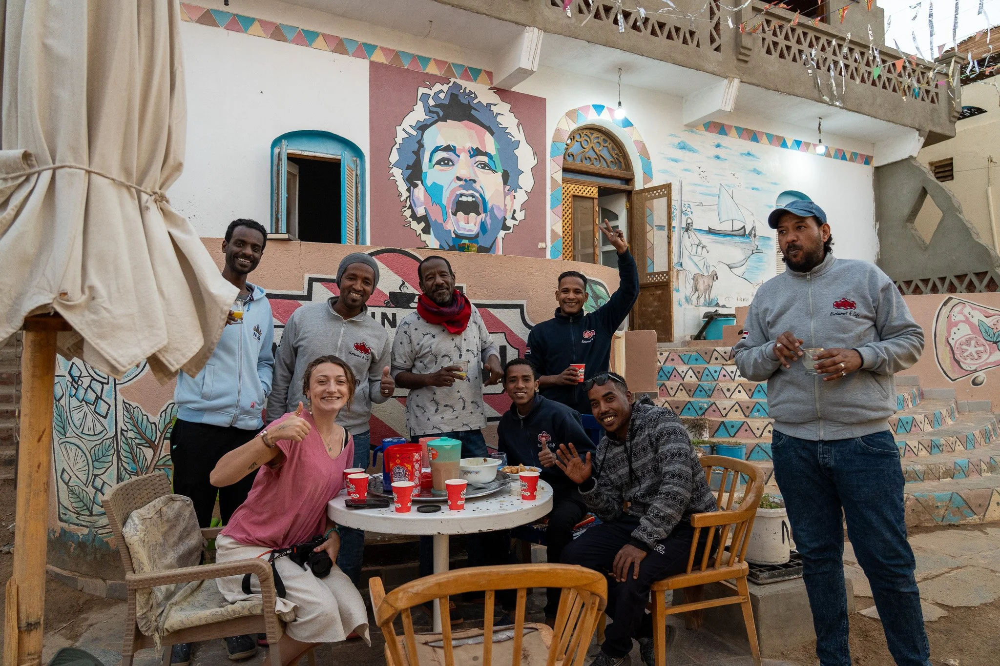 Group of eight people gathered around a table outdoors, smiling and celebrating, with a colorful mural of a woman's face and a sailboat in the background.