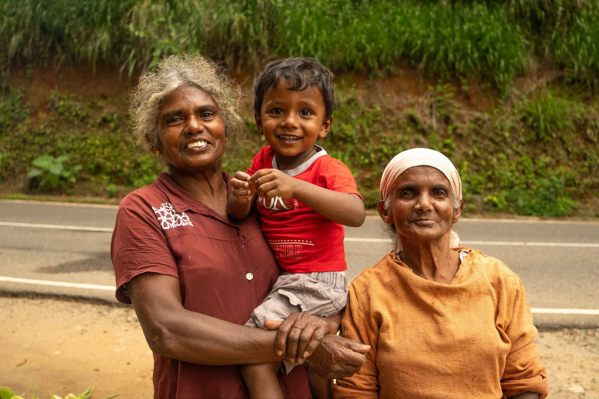 Three people, an elderly woman with a headscarf, another elderly woman, and a young boy, standing outdoors on a roadside with greenery in the background. The woman on the left is carrying the boy on her arm, and all are smiling.