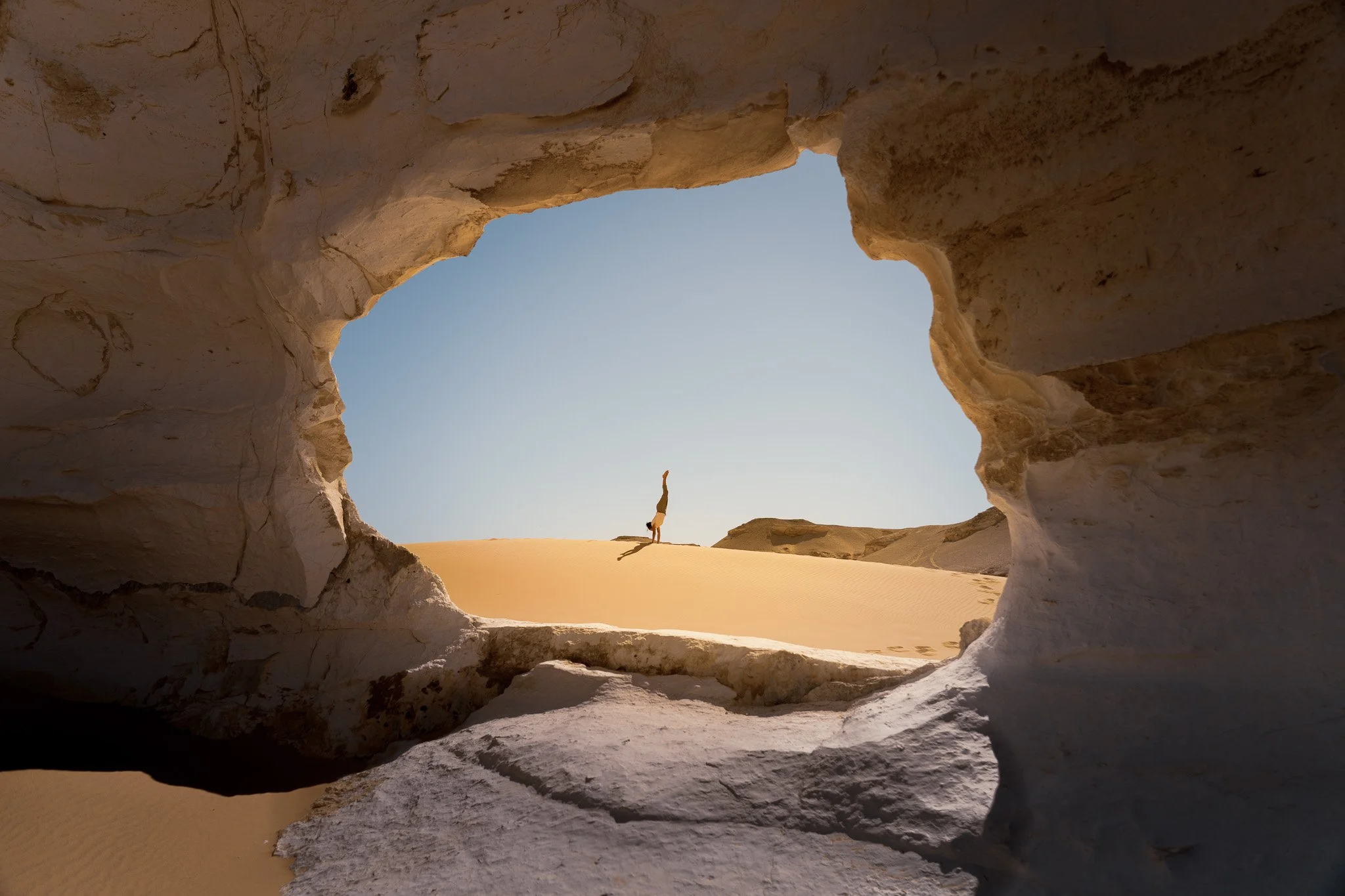 A person doing a handstand on a sand dune, viewed through a large rock formation opening in a desert landscape.