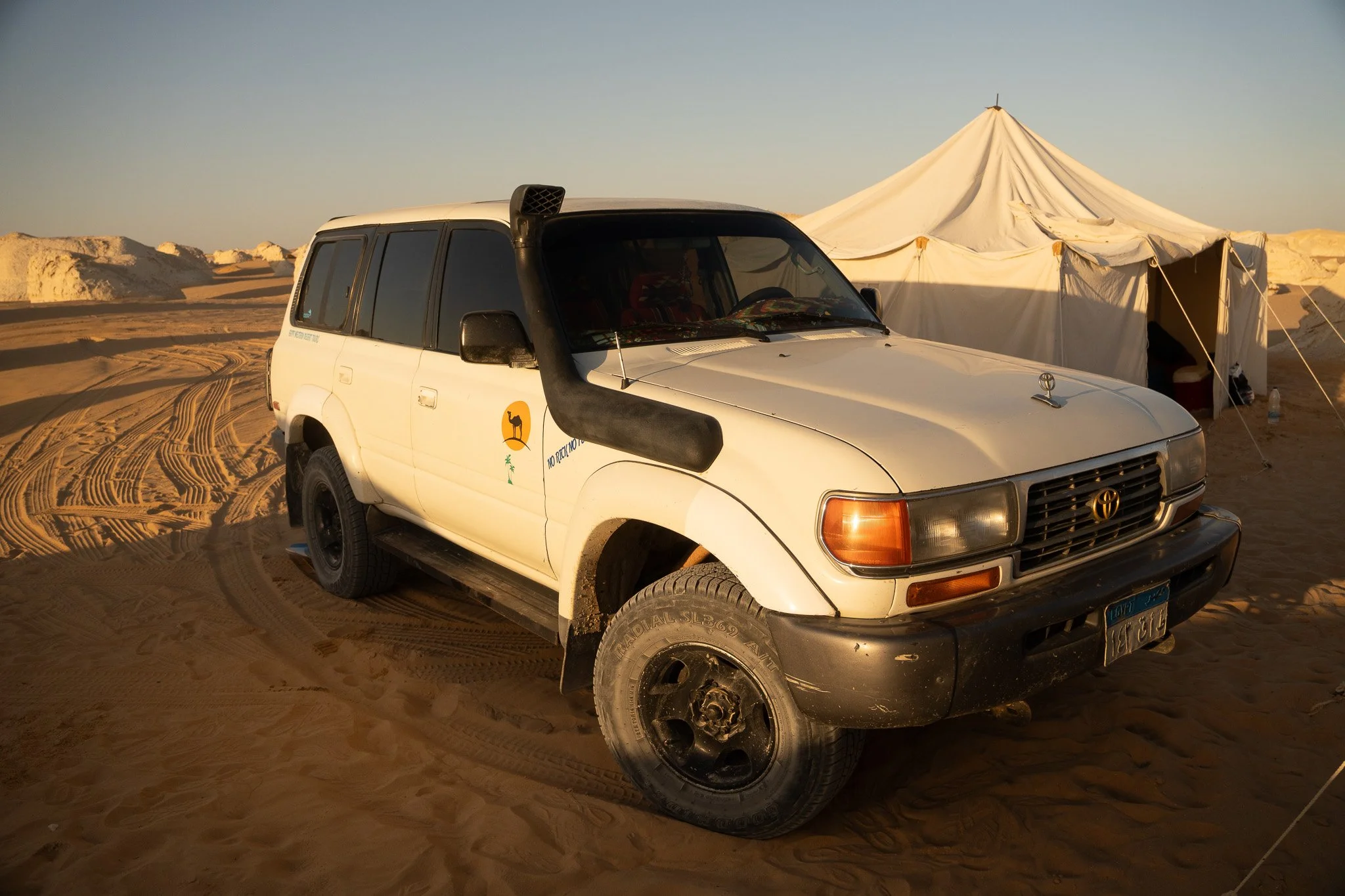A white Toyota SUV parked on desert sand near a white tent with sand dunes in the background.