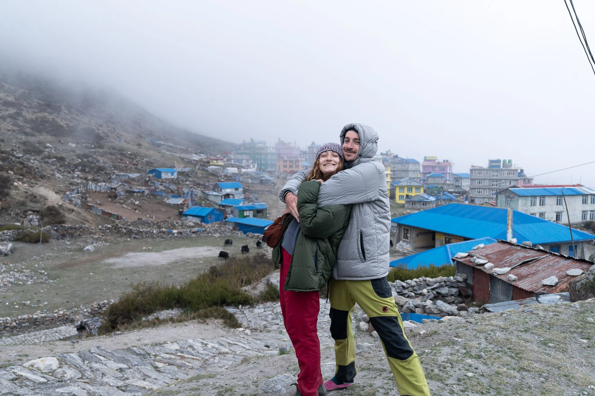 Two people hugging on a hillside with a mountain village in the background. The woman wears a purple hat, green jacket, and red pants while the man wears a gray hoodie, gray jacket, and yellow pants. The village has multicolored buildings with blue r