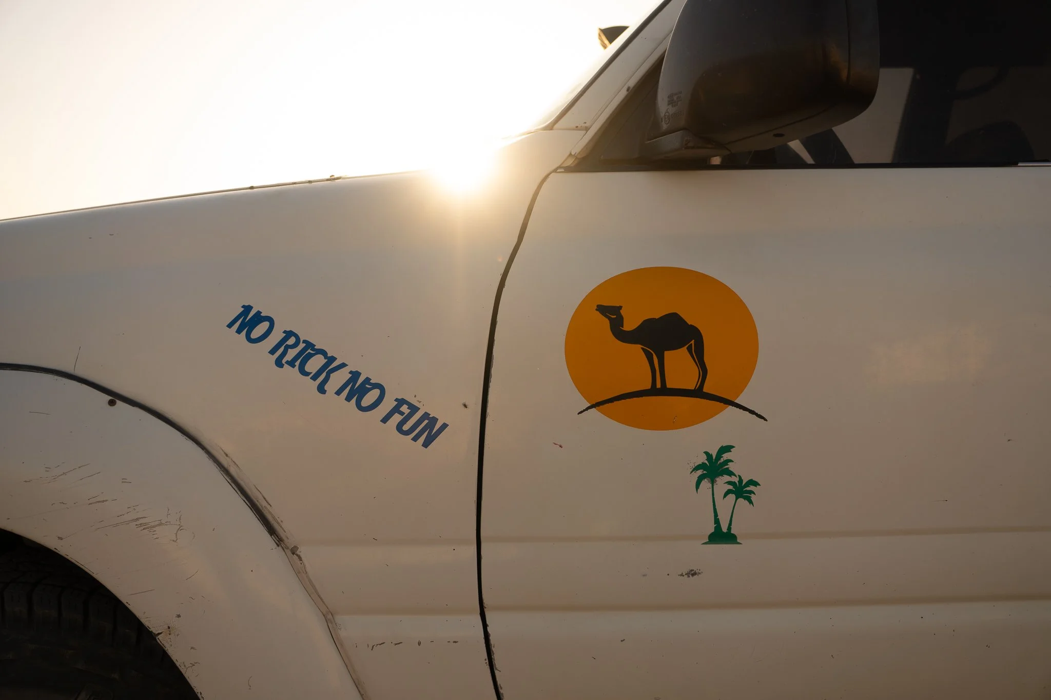 Close-up of a white vehicle with a camel and palm tree stickers, and the phrase "NO RICK NO FUN" written on the side, with the sun shining behind the vehicle.