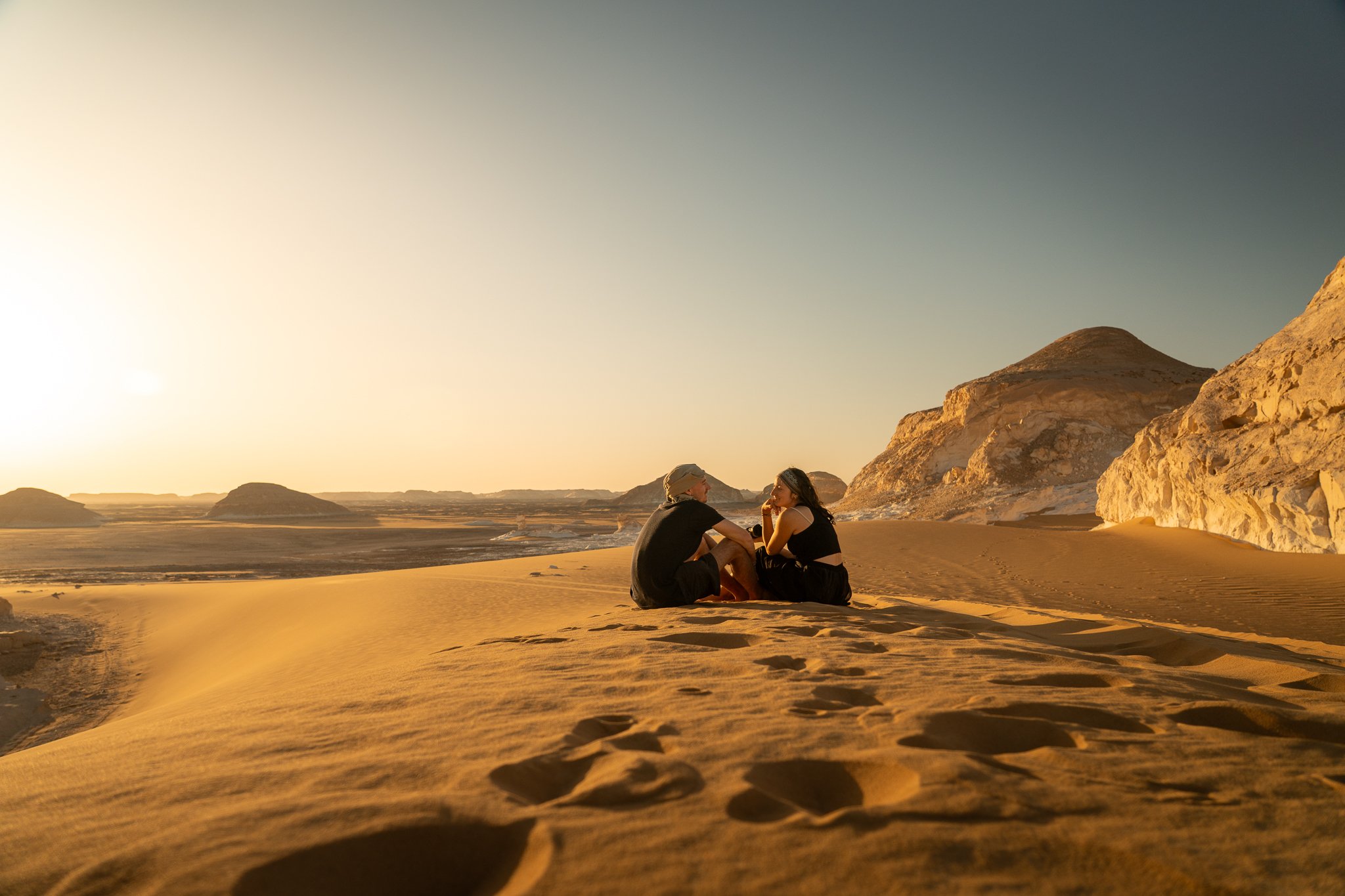 A man and woman sitting on desert sand dunes, facing each other and talking, with mountains in the background under a clear sky at sunset.