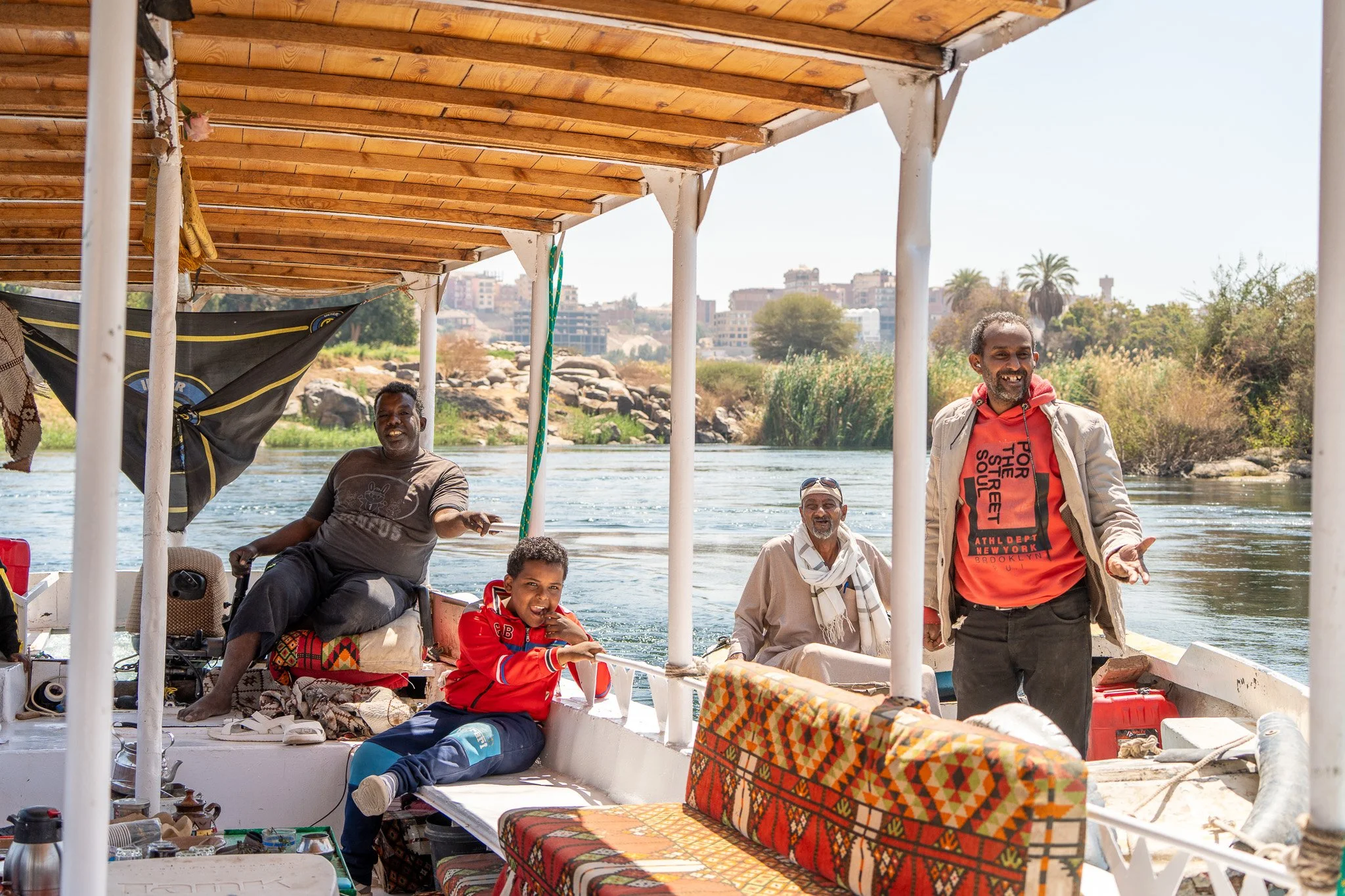 Five men and a boy on a boat smiling and enjoying a boat ride on a river with a cityscape, trees, and rocks in the background.