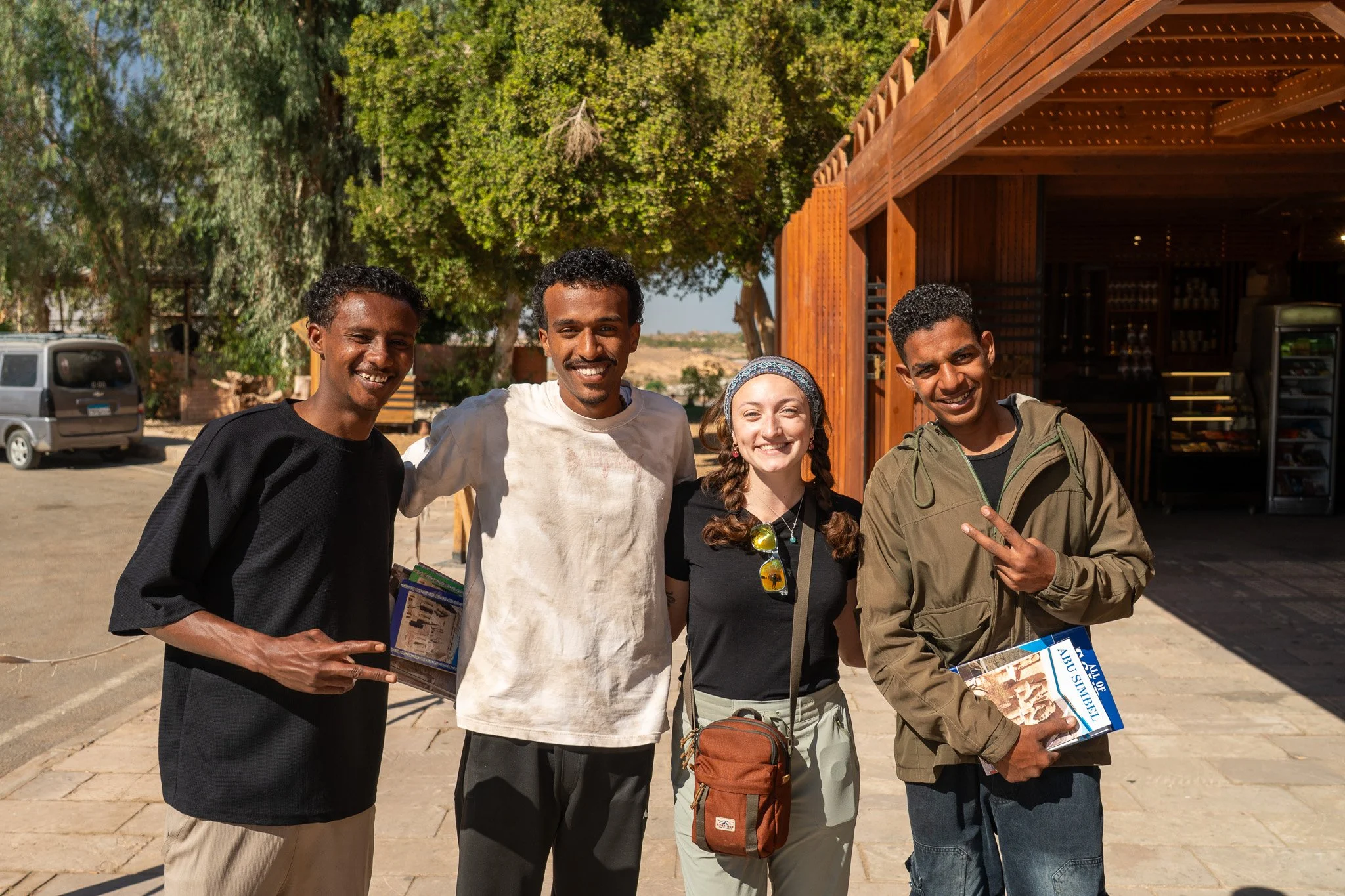Four friends smiling and posing outdoors, with two men making peace signs, one woman carrying a small bag, and a man holding a magazine, standing in front of a wooden building with trees and vehicles in the background.