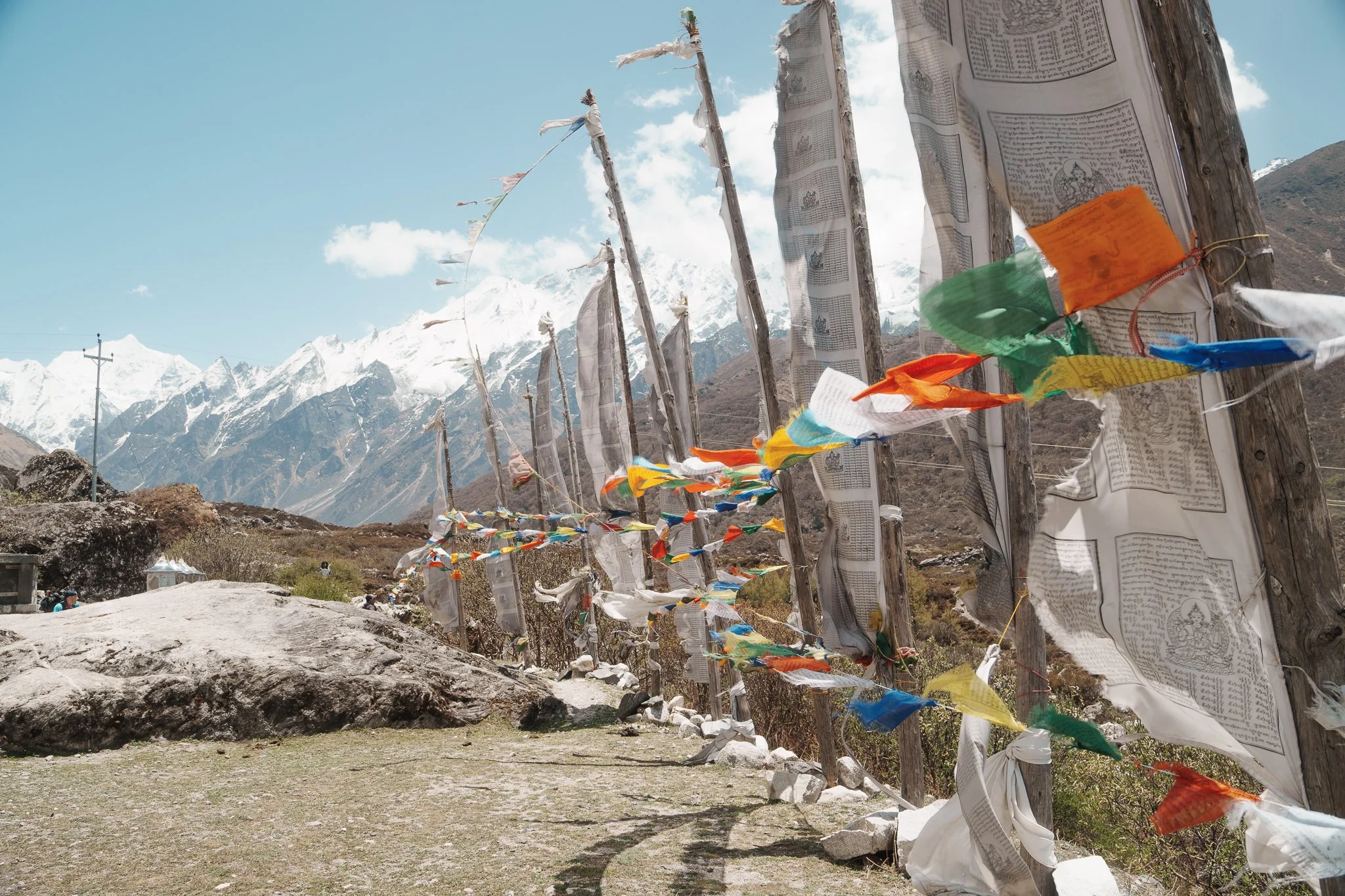 Prayer flags on wooden poles along a mountain trail with snow-capped peaks in the background.