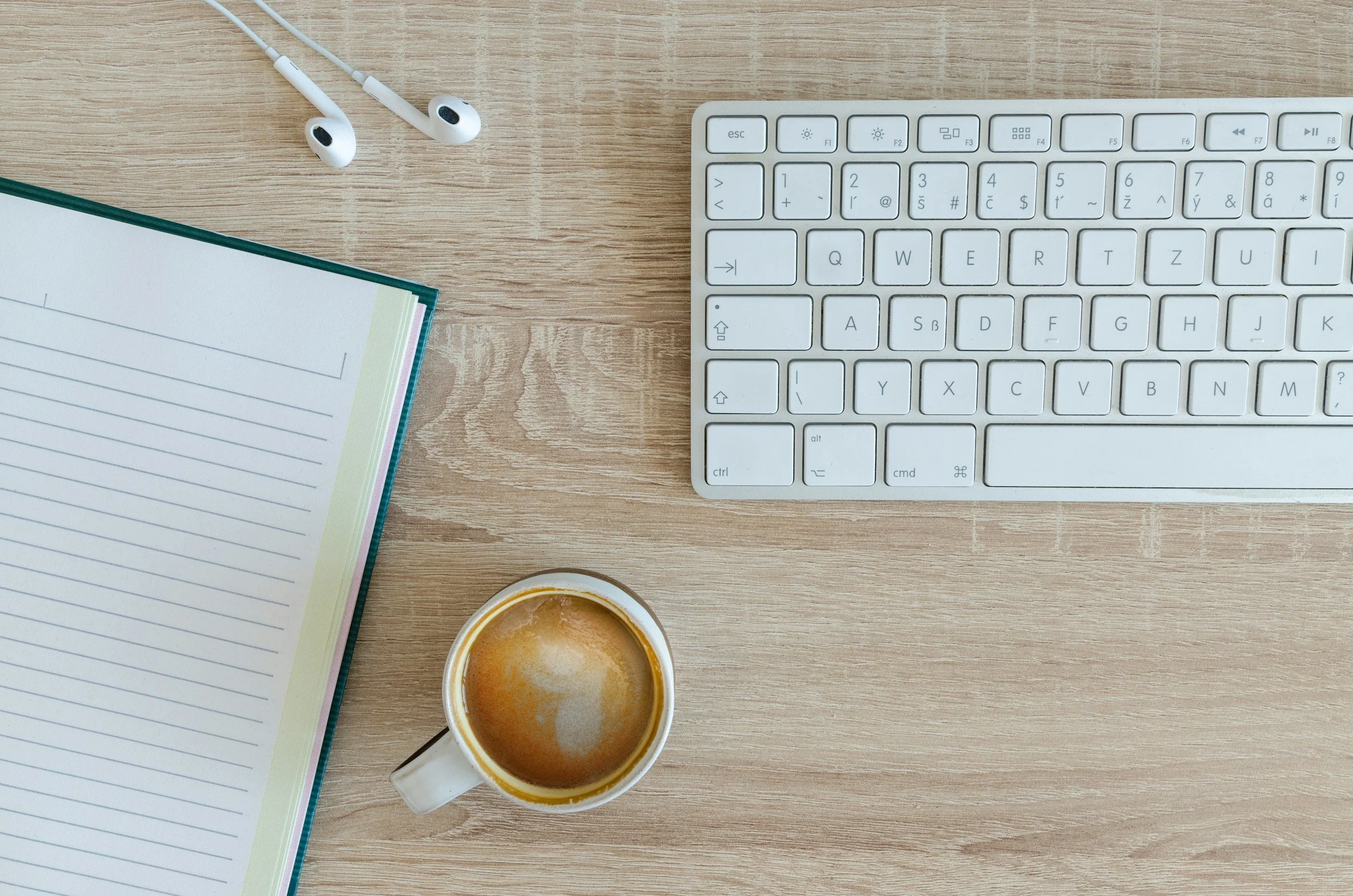 A top-down view of a workspace with a notebook, a cup of coffee, a white keyboard, and a pair of white earphones on a wooden surface.