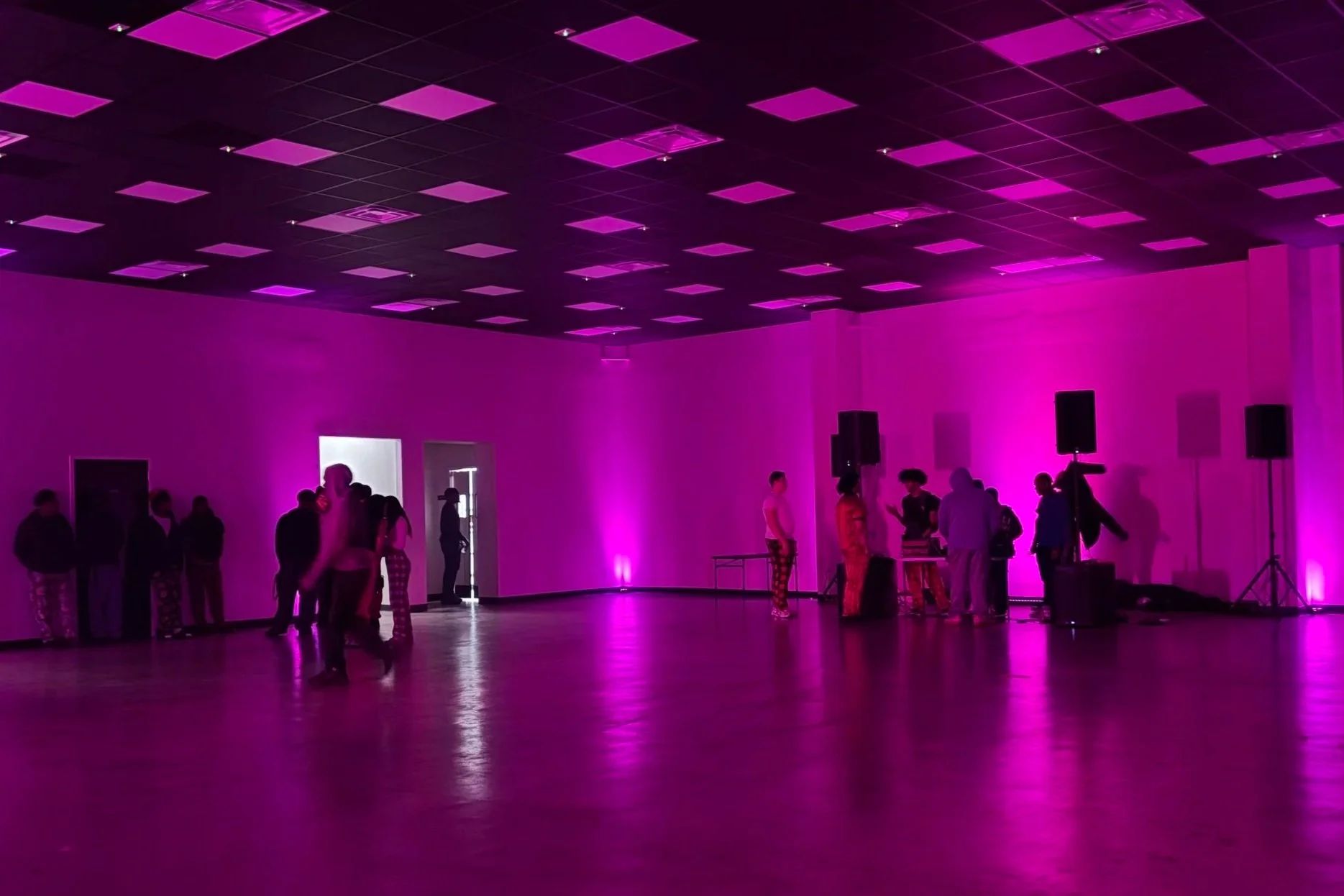 People gathering in a dimly lit room illuminated in purple with a reflective floor, speakers, and a DJ setup.