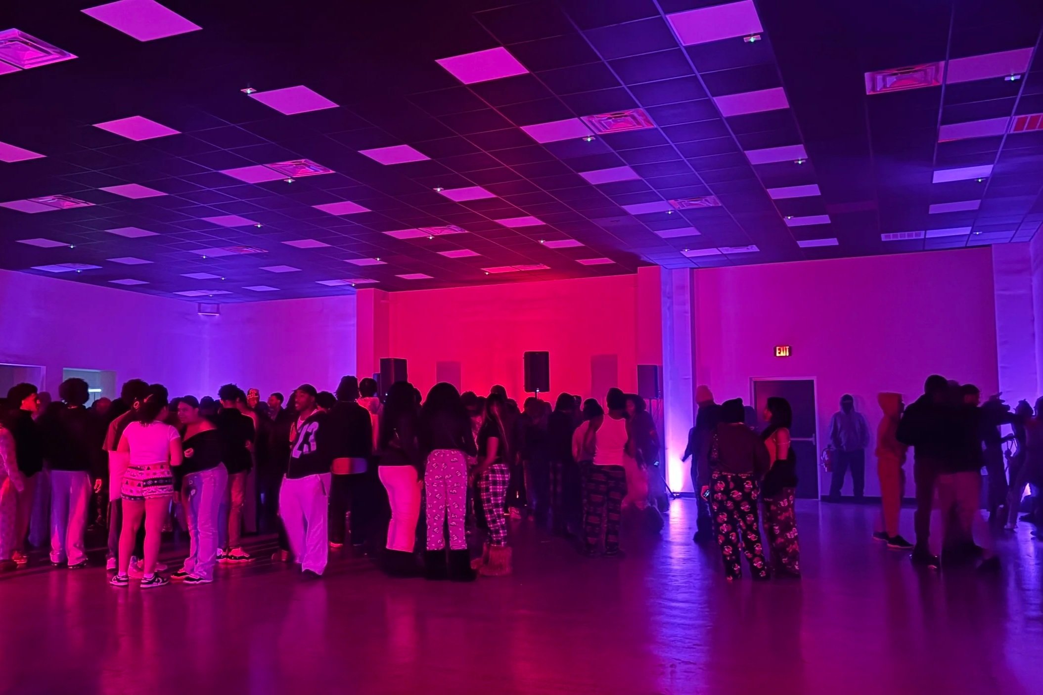 People gathered at an indoor event with vibrant purple and pink lighting, possibly a dance or party, in a large room with a high ceiling.