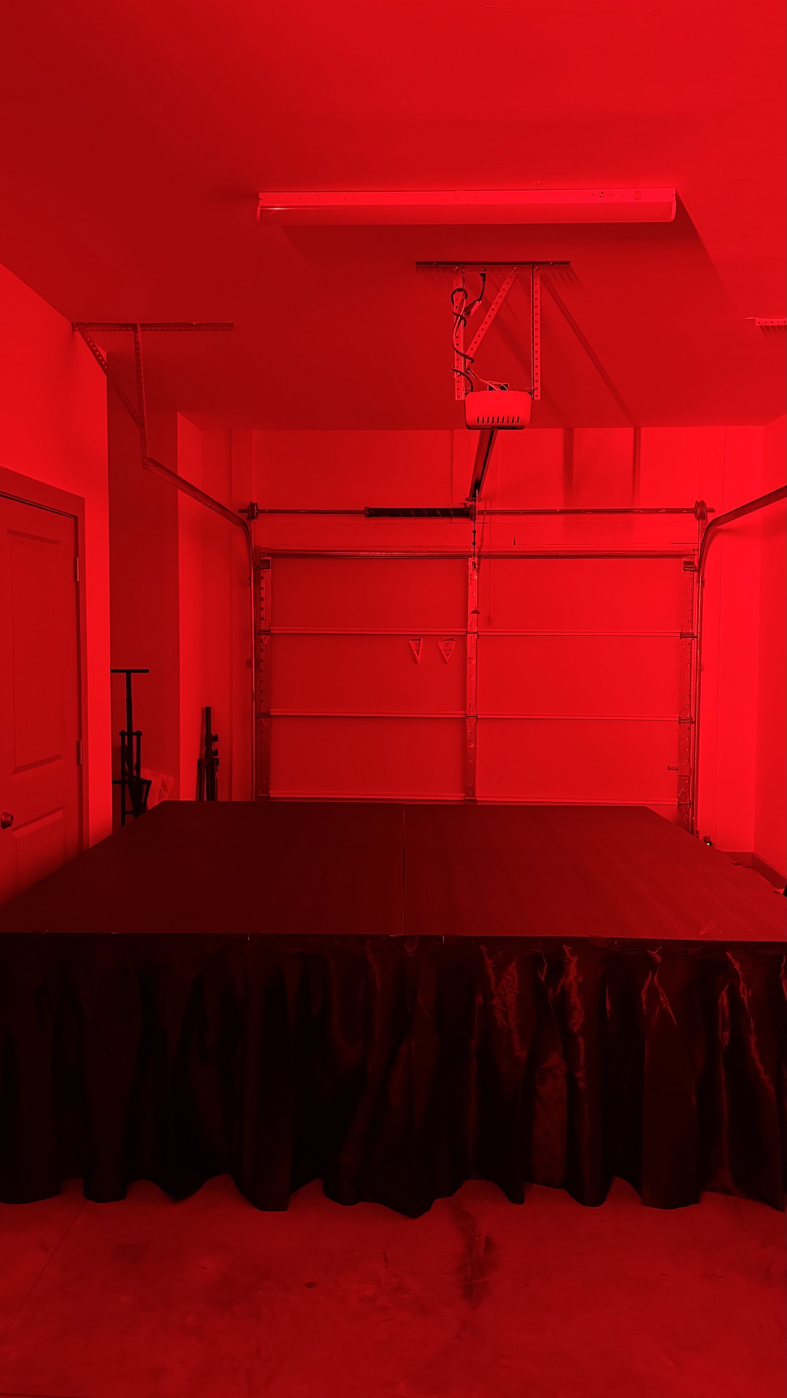 A garage with a raised ceiling, a closed garage door, and red lighting. There is a black table or platform with a black tablecloth in the center of the room, and some items on the left side near a door.