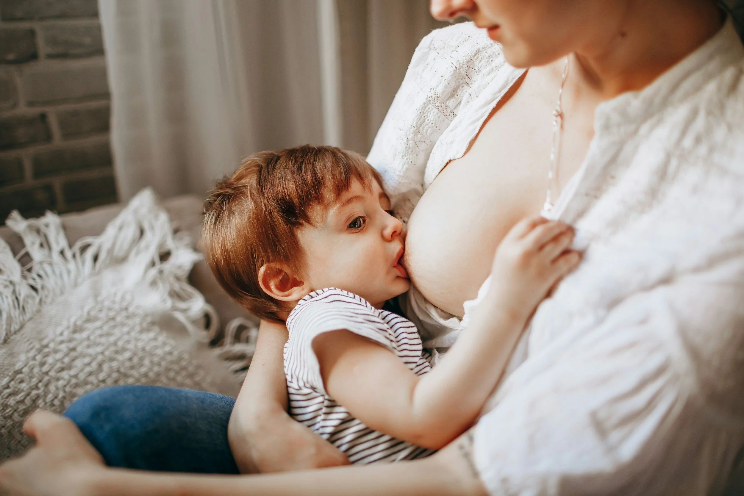 A young child with red hair breastfeeding from a woman, possibly his mother, in a cozy indoor setting.