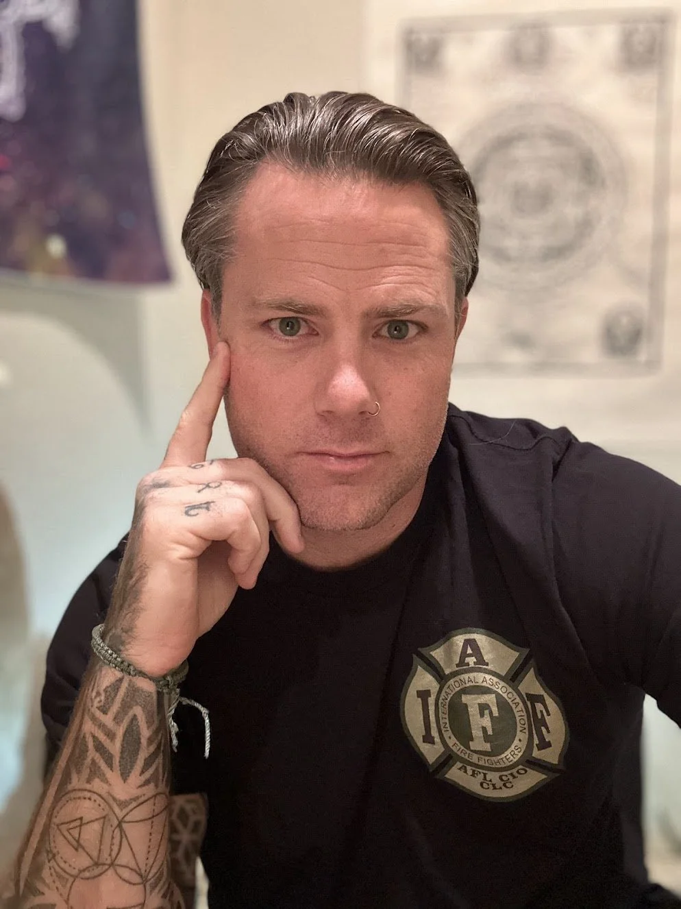A man with tattoos on his arm and a nose ring, resting his chin on his hand, wearing a black shirt with a firefighter emblem, in a room with abstract black and white wall art and a lamp in the background.