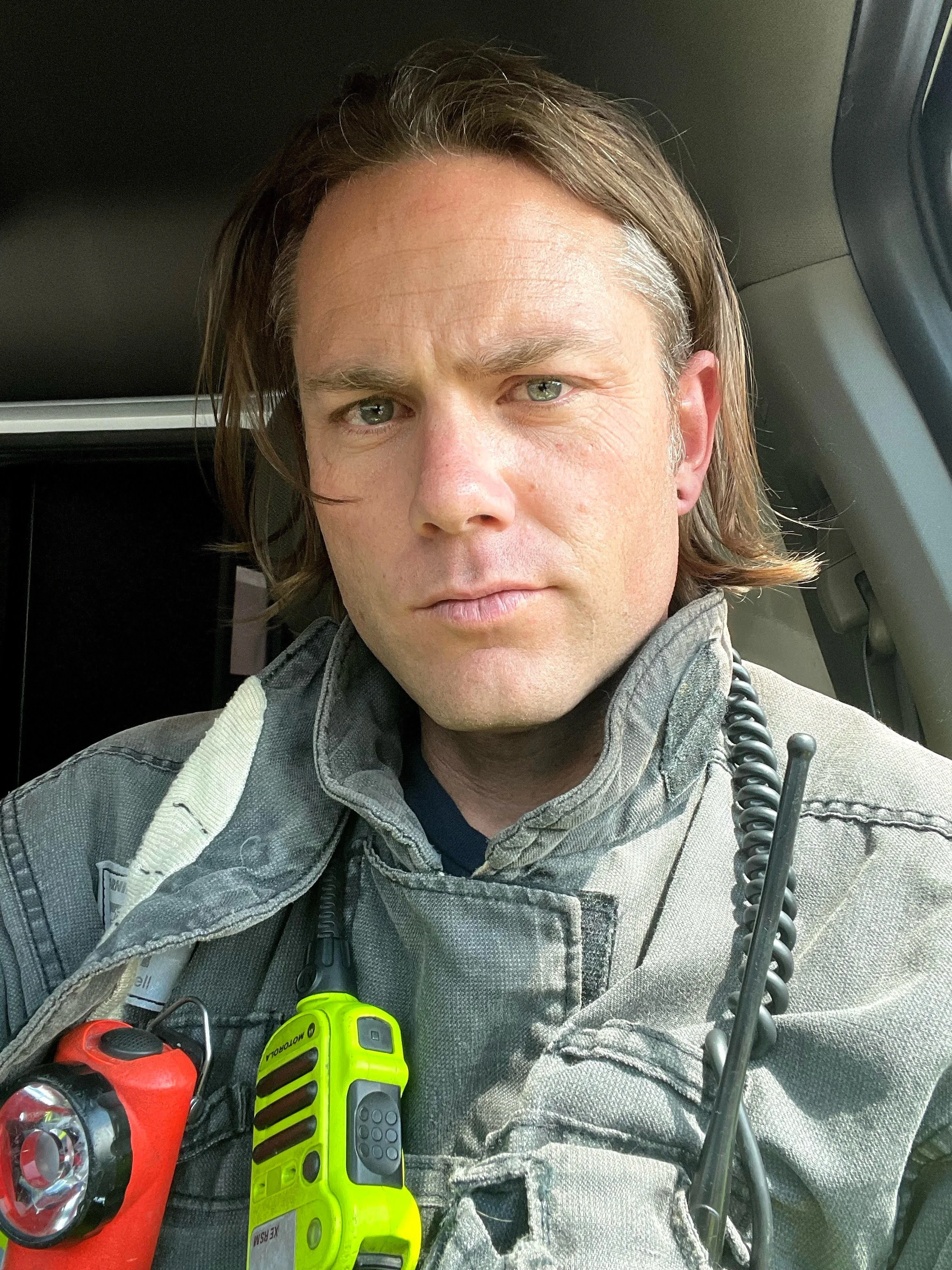 A man in a firefighter uniform sitting in a vehicle, with a headlamp and a walkie-talkie attached to his uniform.