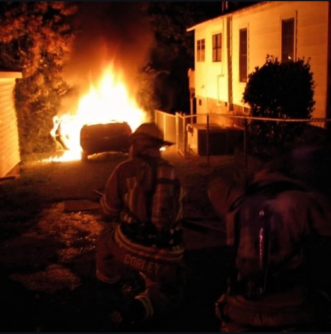 Two firefighters crouch near a burning vehicle in a backyard at night.