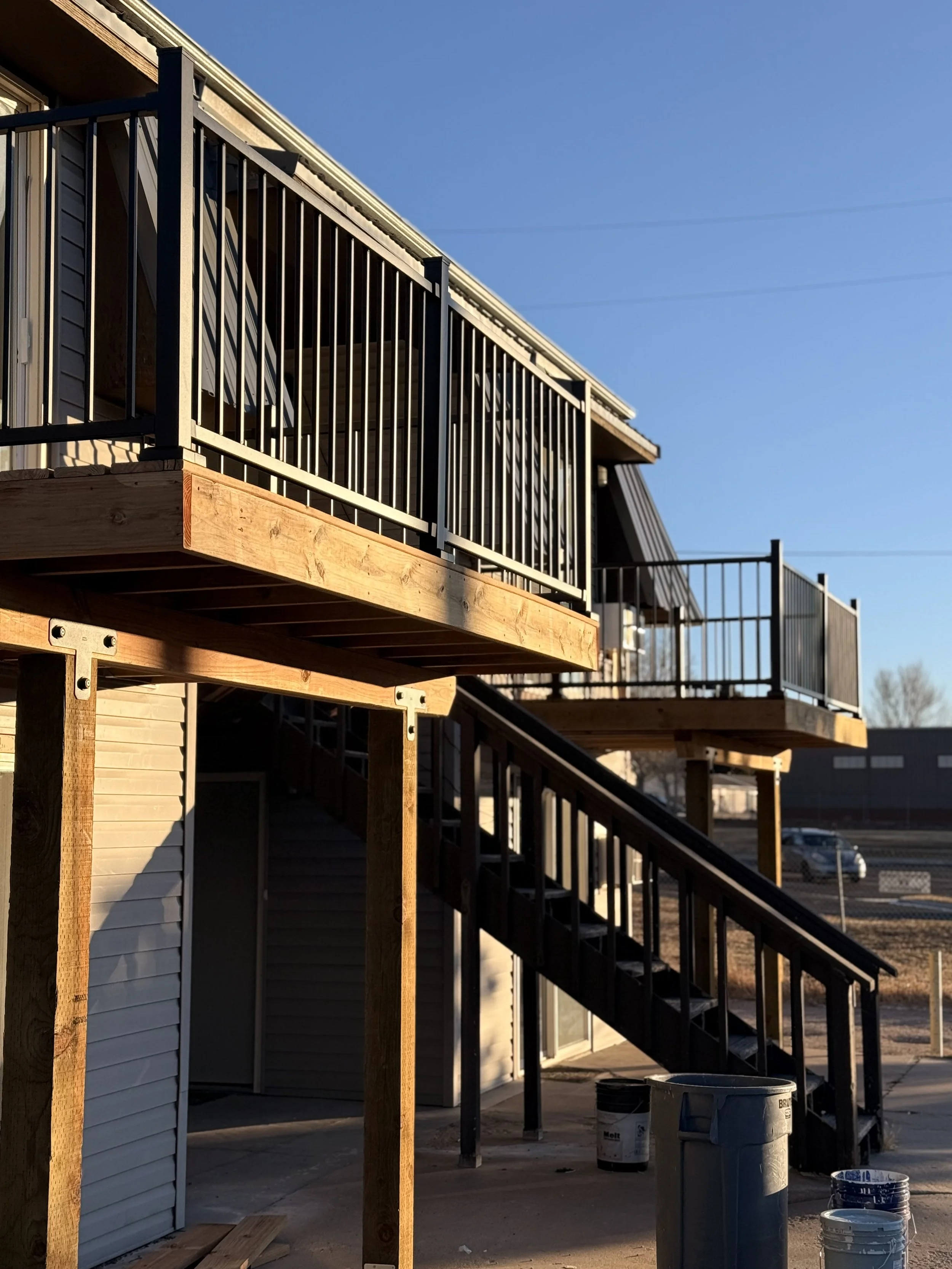 A newly constructed wooden patio with black metal railings attached to a residential building, with stairs leading down to the ground. Construction supplies are visible at the base of the stairs.