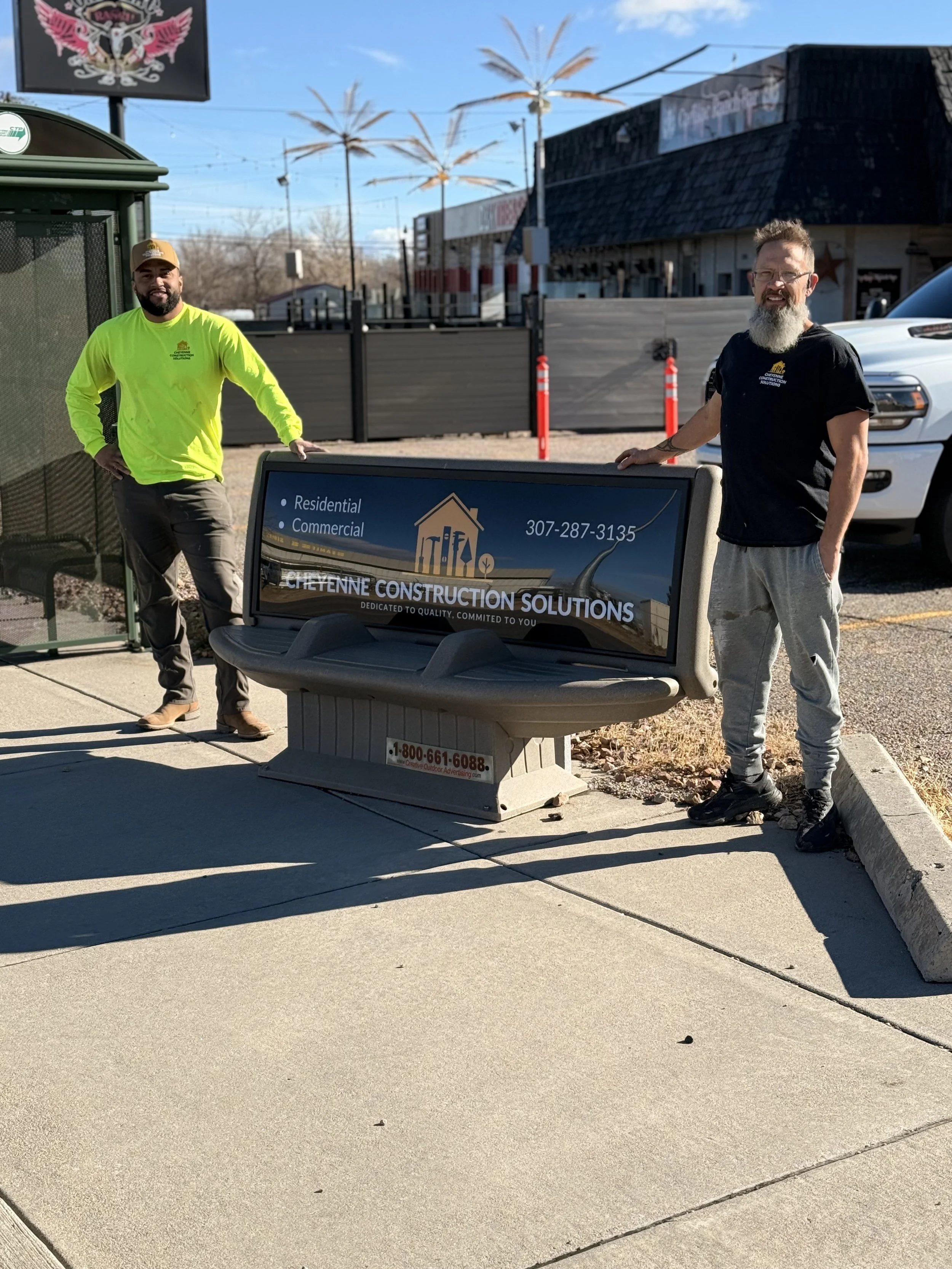 The two owners standing next to a bench with advertisement for Cheyenne Construction Solutions, in an outdoor area with palm trees, a building, and a parking lot.
