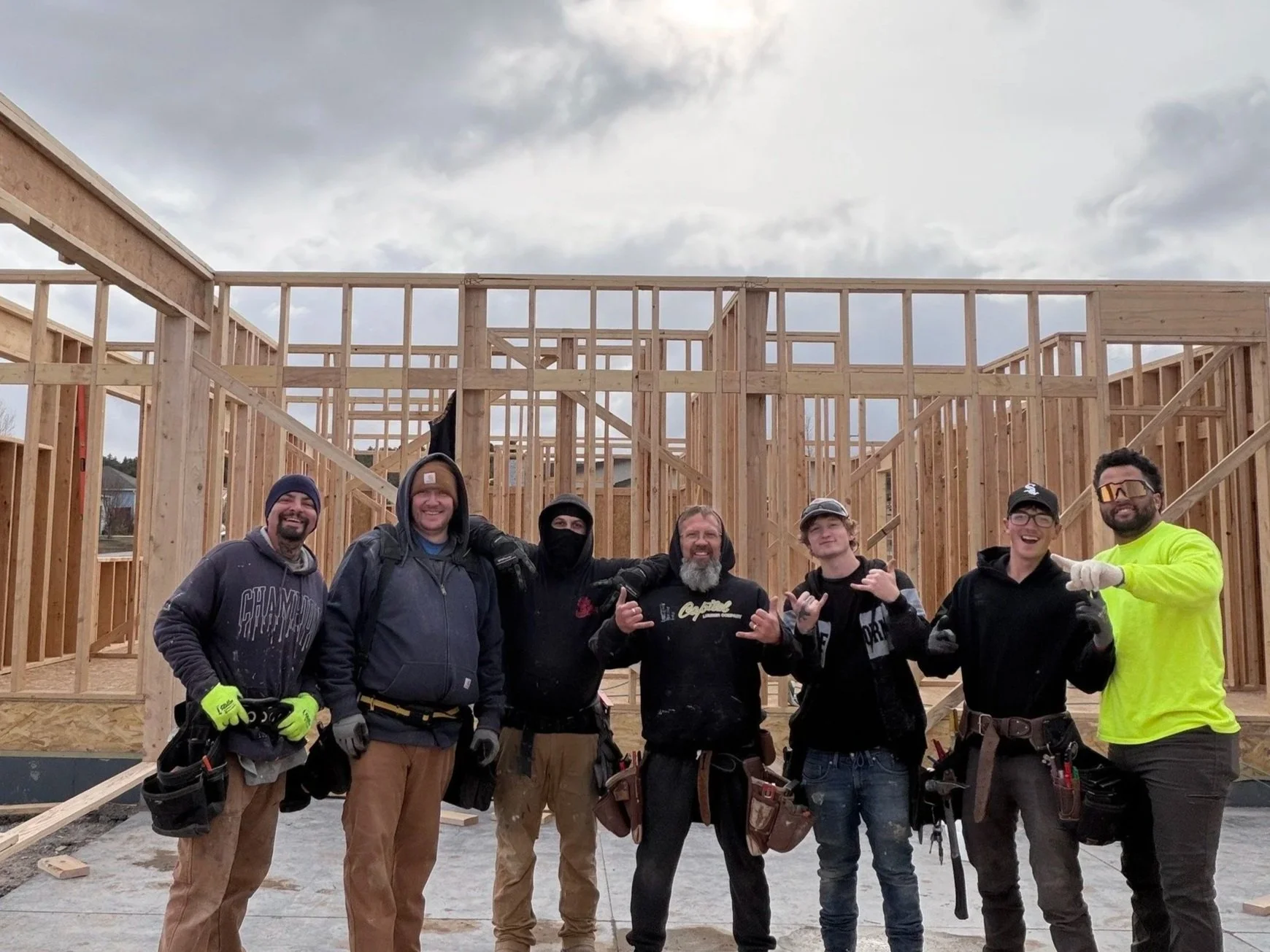 Group of seven construction workers smiling and posing in front of a wooden framing structure during construction.