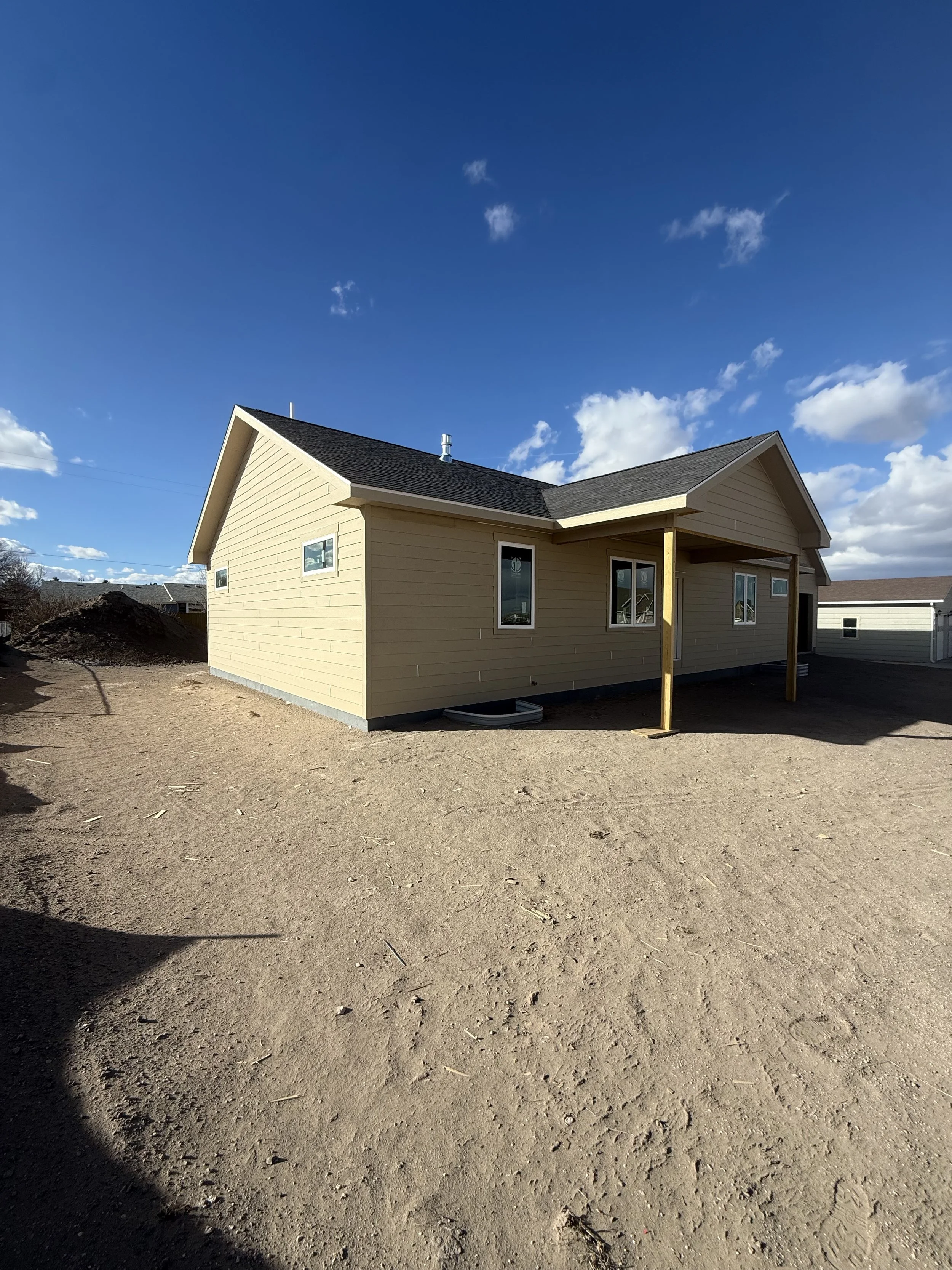 Newly built beige house with black roof and small covered porch under construction, set against a blue sky with scattered clouds, dirt yard in foreground.