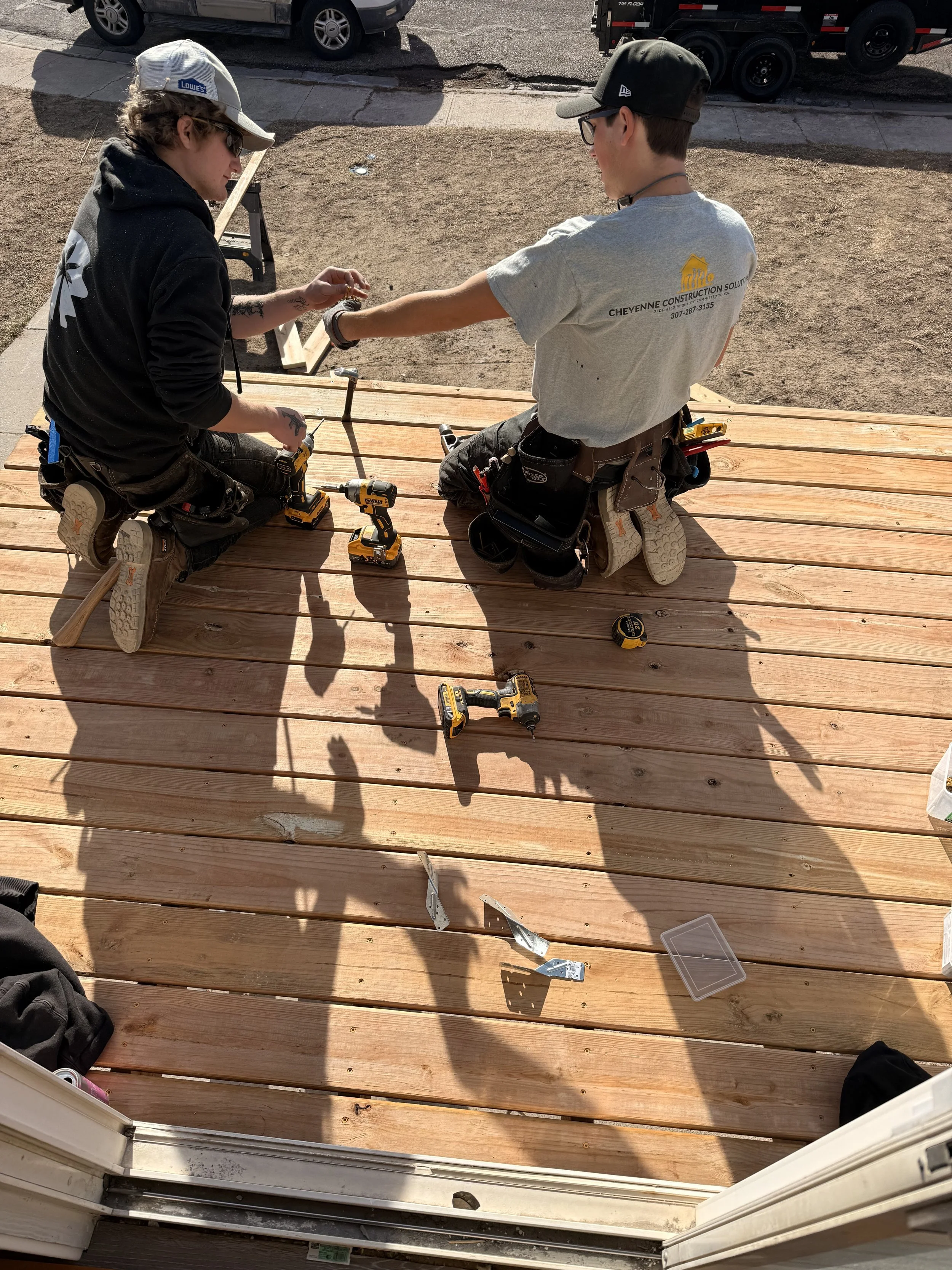 Two construction workers kneeling on a wooden deck, working together with power tools, surrounded by construction materials and tools.