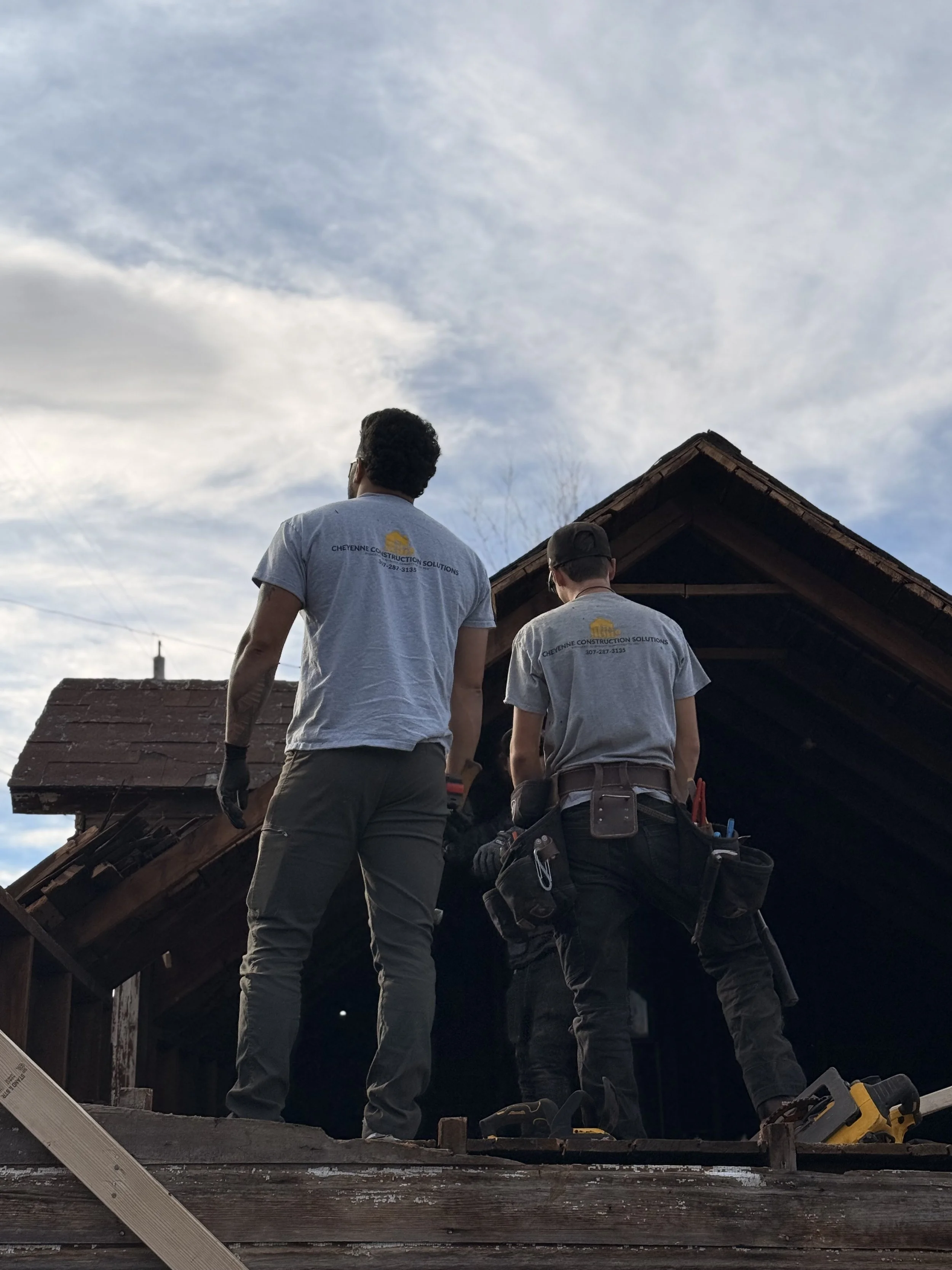 Two construction workers standing on the roof of a house, working on repairs or renovation, with a partly cloudy sky in the background.