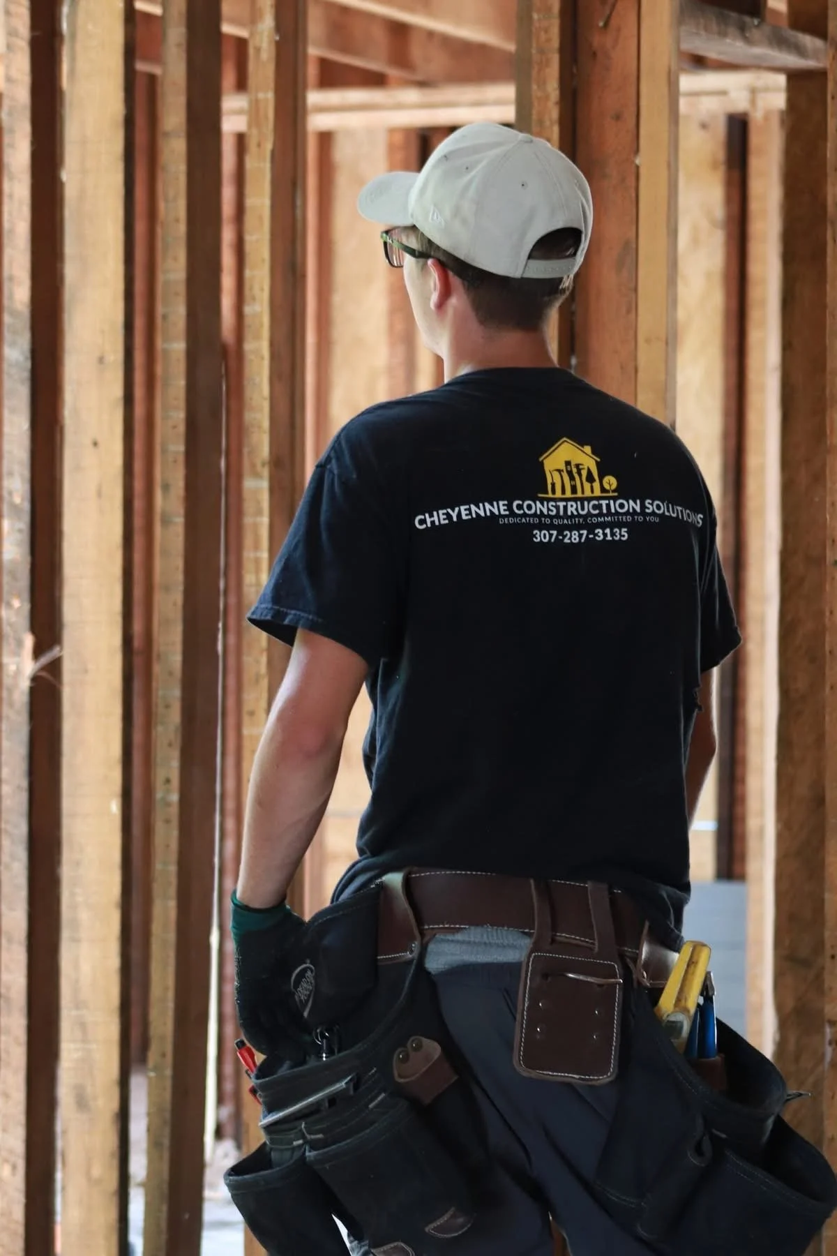 A construction worker wearing a black shirt with 'Cheyenne Construction Solutions' on the back, a white cap, and gloves stands inside a wooden framing structure. The worker's tool belt is equipped with various tools.