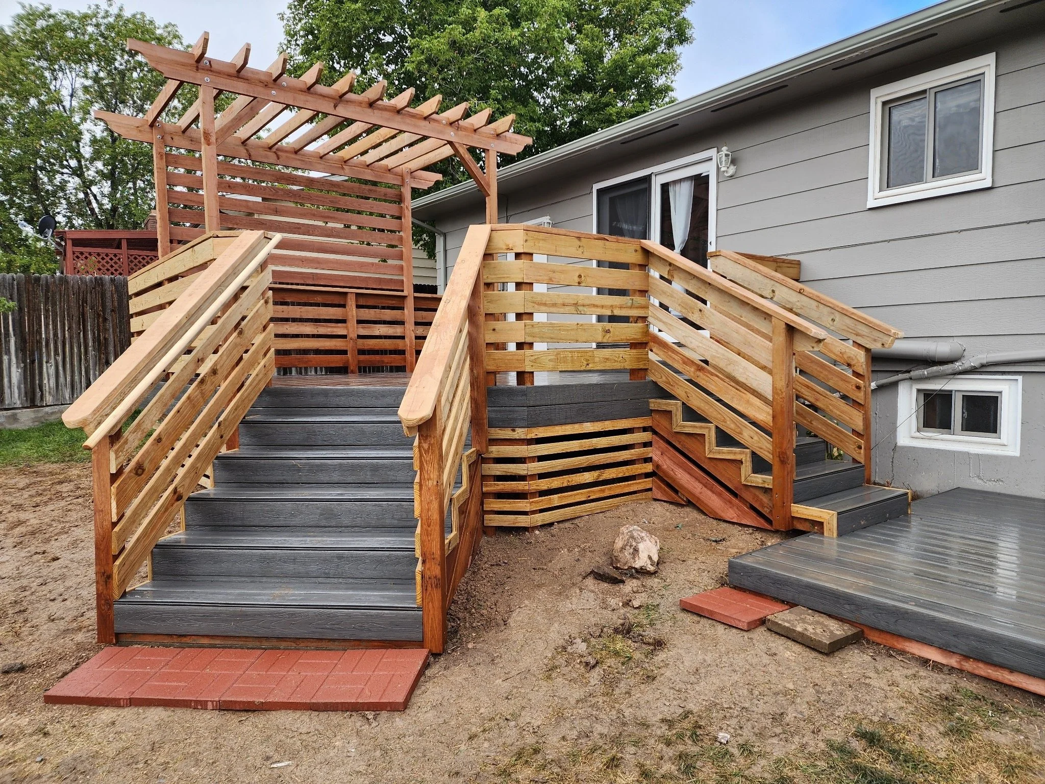 Exterior of a two-story building with a wooden staircase and balconies with black metal railings, construction materials on the ground, under a clear blue sky.