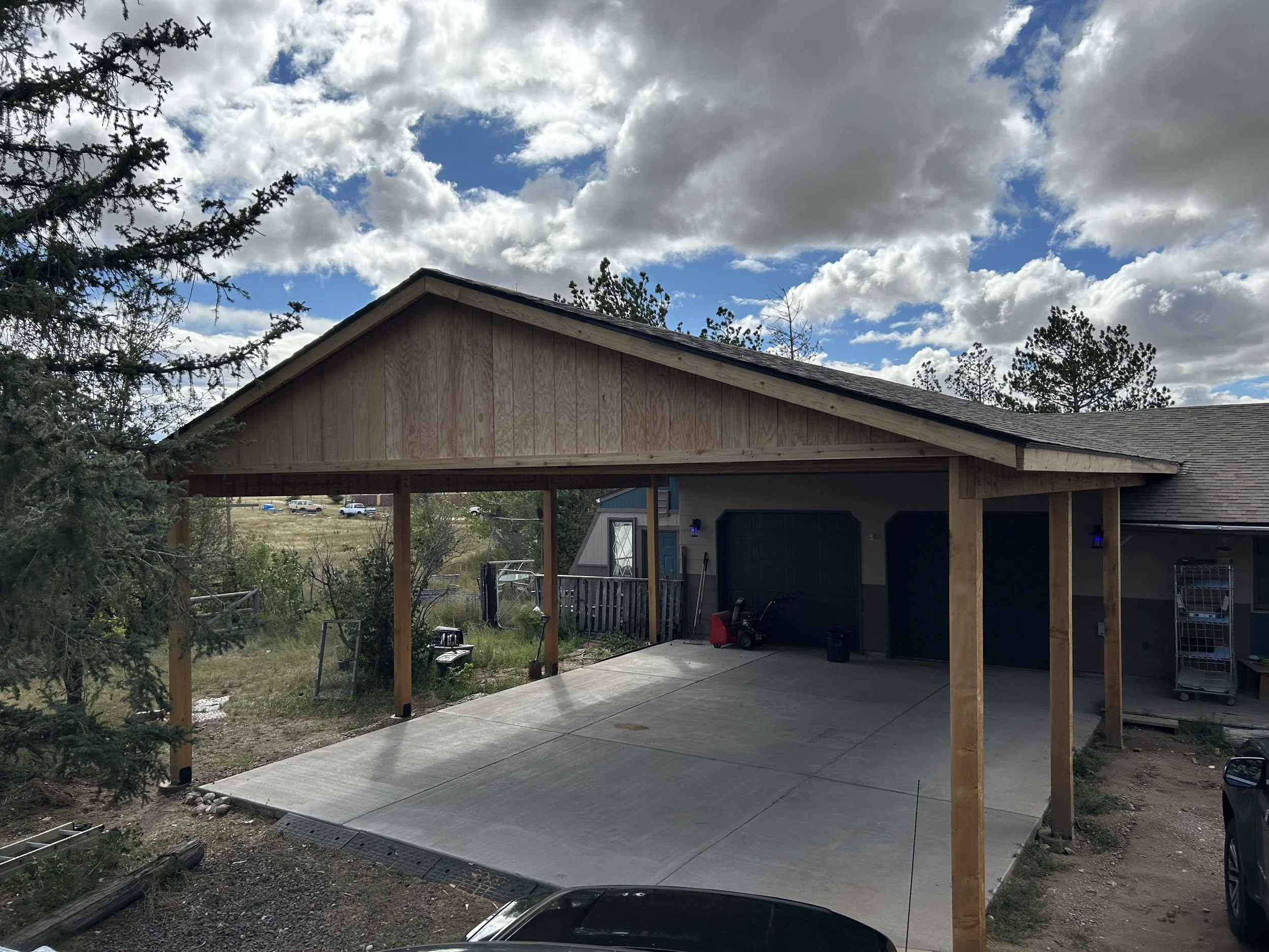 A newly constructed wooden carport attached to a house, with a concrete driveway, some gardening tools, and a cloudy sky overhead.