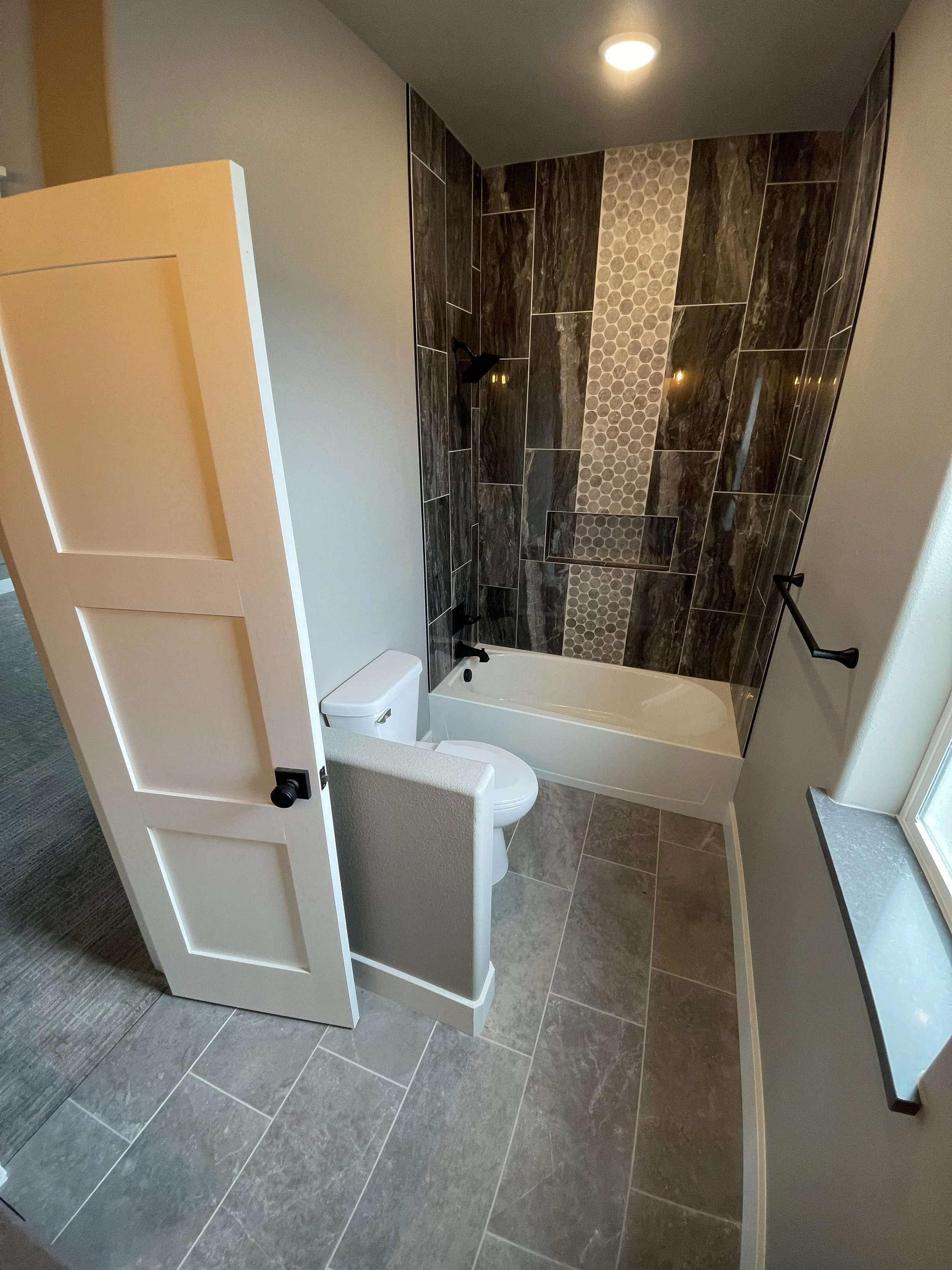 Small bathroom with a white door, toilet, bathtub with dark tile walls featuring a vertical strip of small hexagon tile pattern, window with a ledge, and a black towel bar.