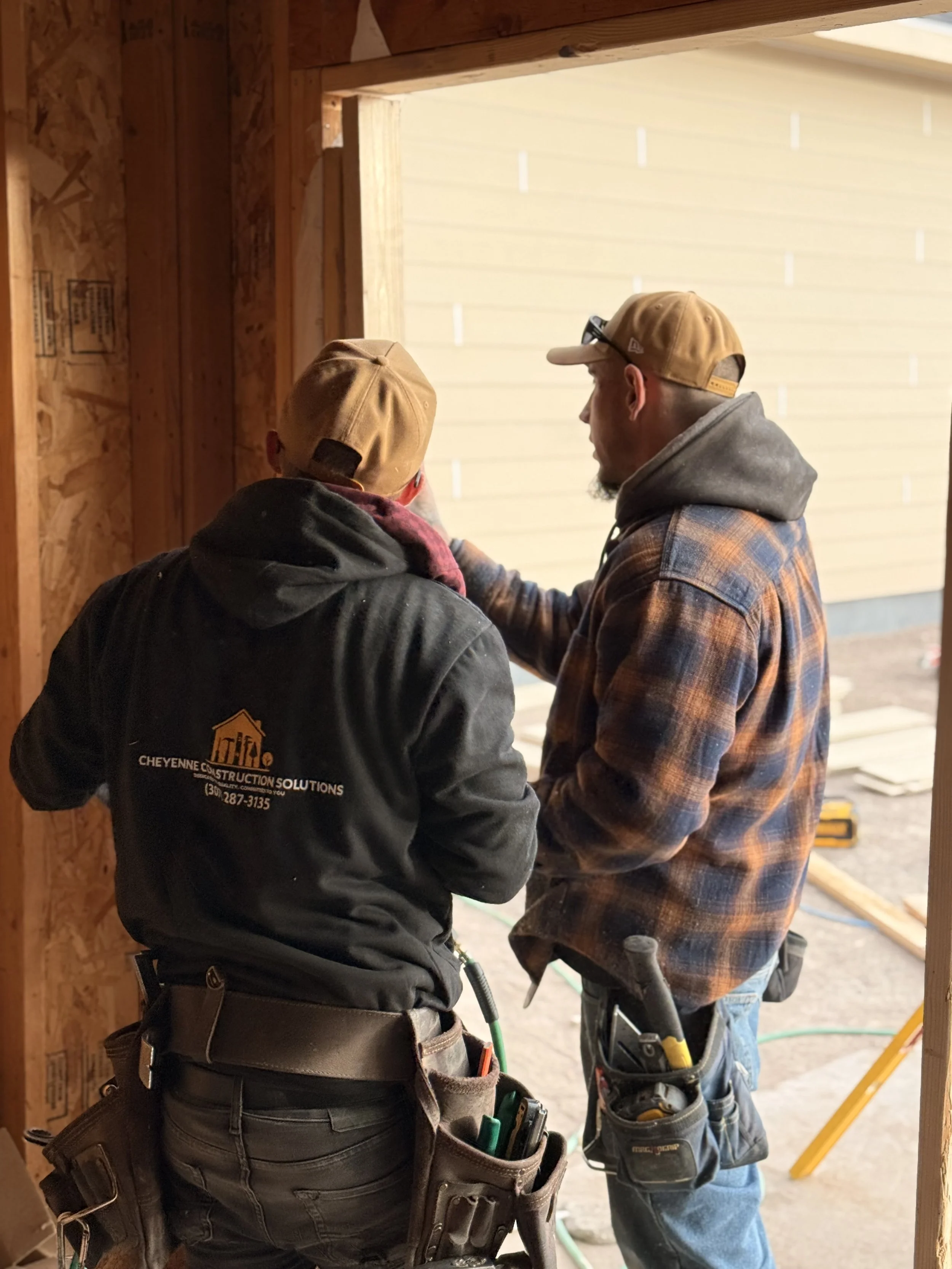 Two construction workers wearing brown hats and work clothes are working together inside a partially built wooden structure, discussing something near an opening leading outside.