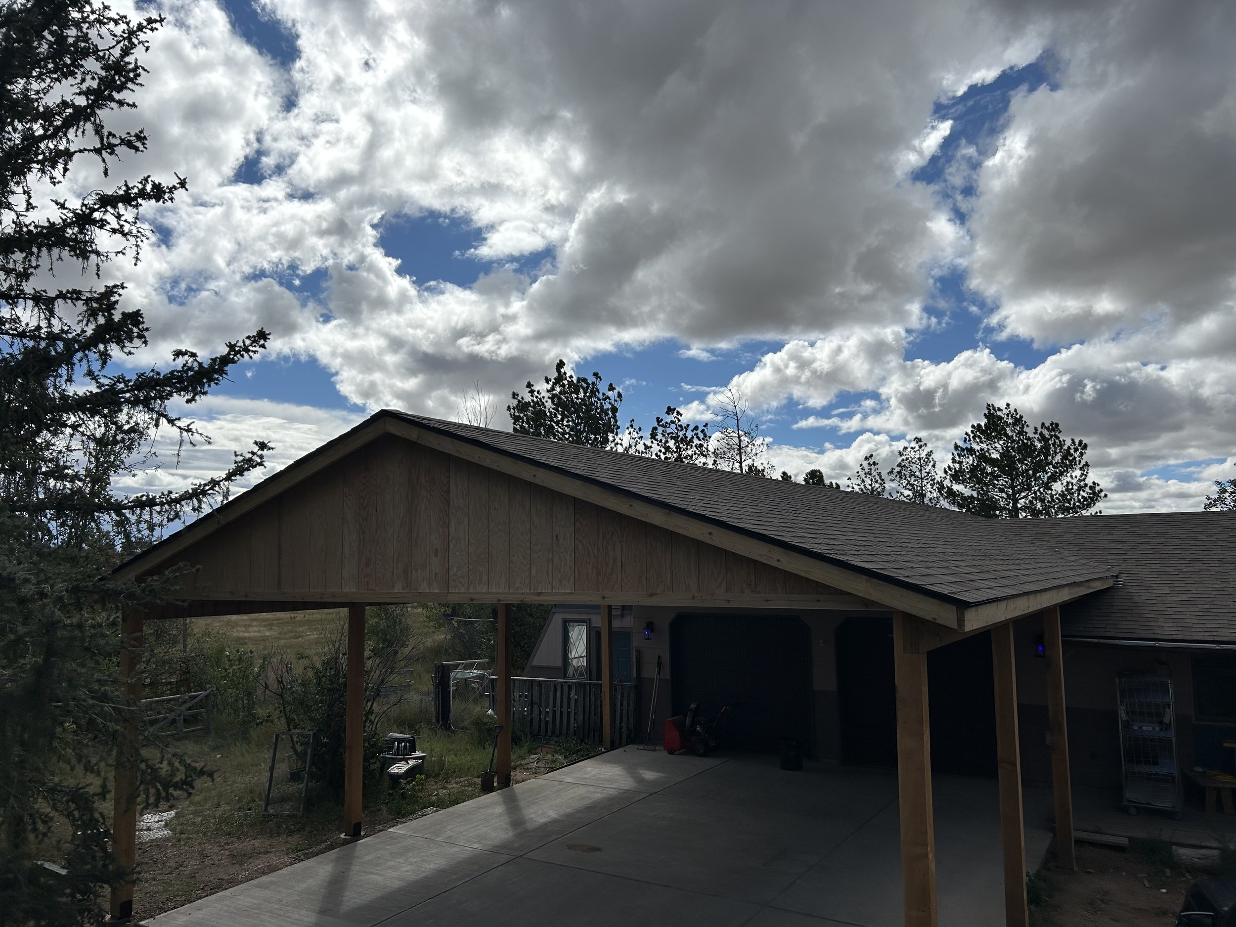Residential house with a wooden carport, cloudy sky with patches of blue, trees in the background, and a concrete driveway.