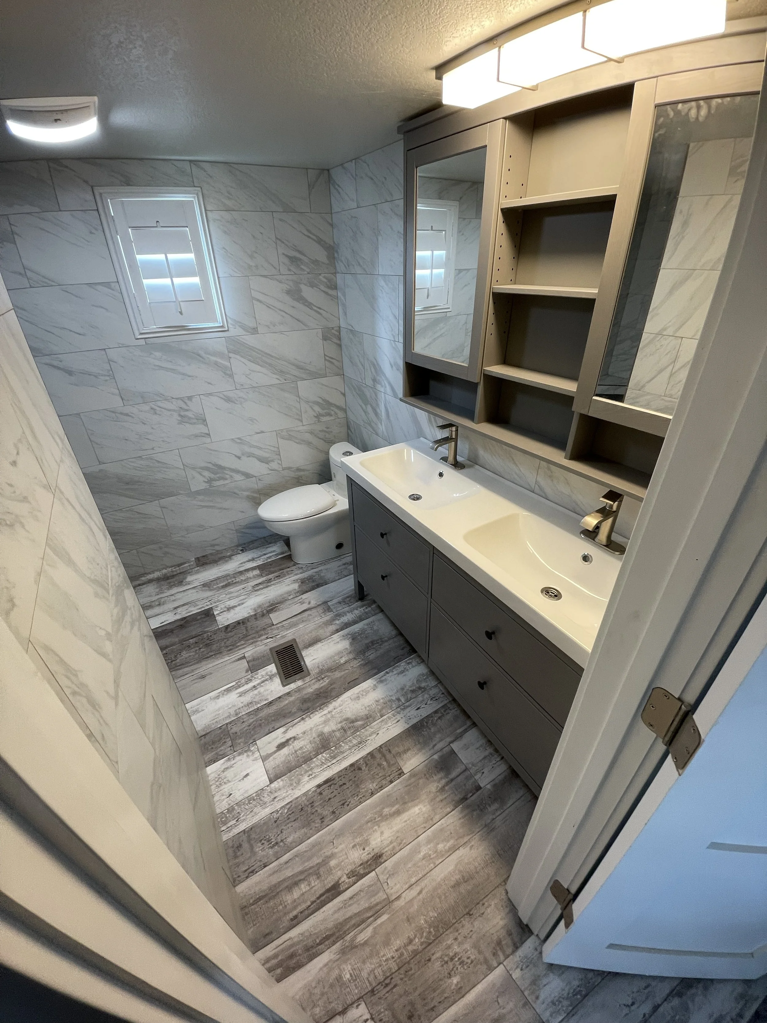 Bathroom with a dual-sink vanity, a large mirror cabinet, marble wall tiles, wood-look tile flooring, a small window, and a toilet.