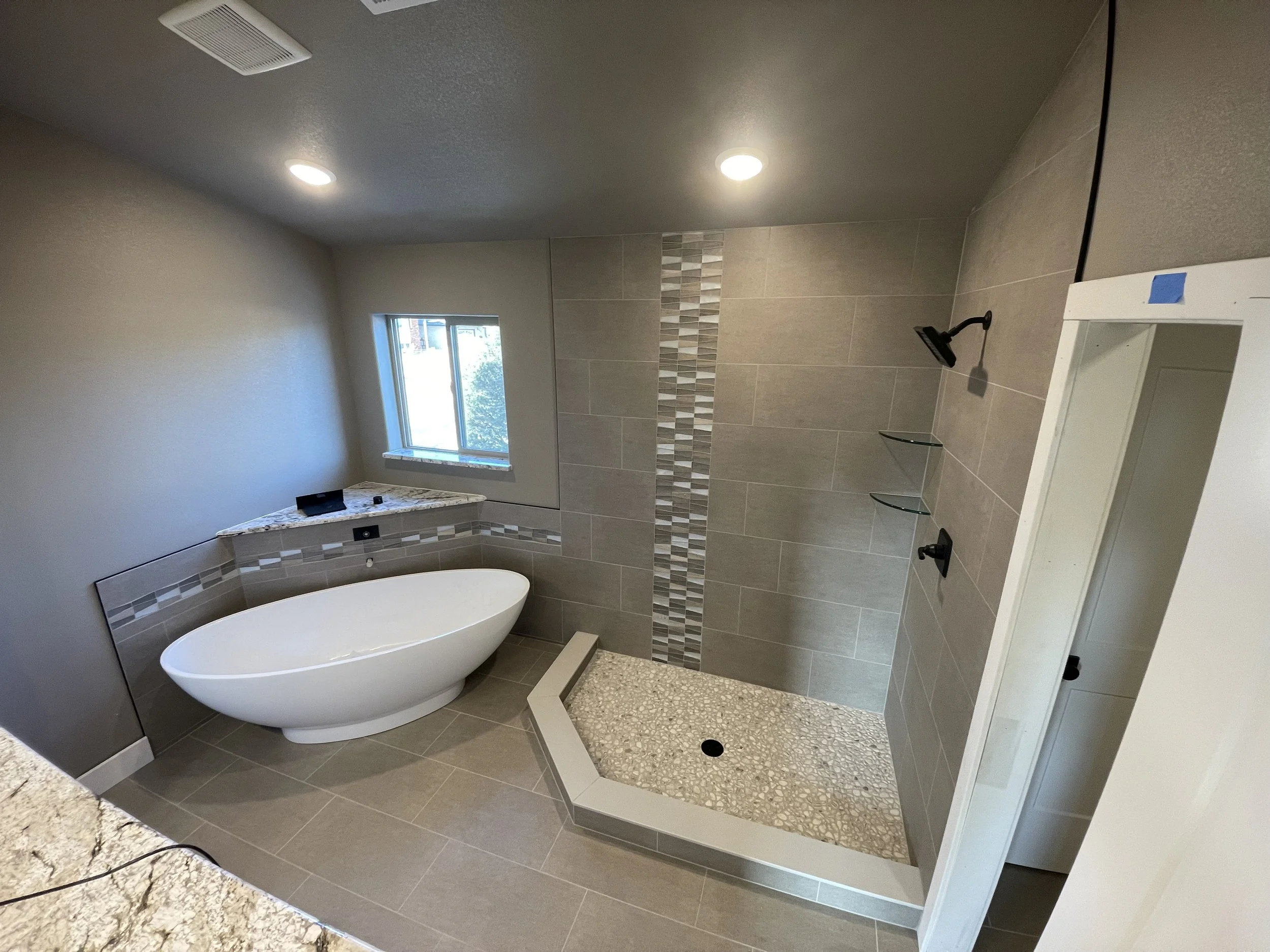 Modern bathroom with freestanding bathtub, walk-in shower with stone tile floor, decorative tile strip, gray wall tiles, small window, ceiling lights, and built-in shelves.