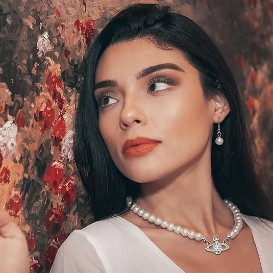 A woman with long black hair, wearing pearl jewelry, and a white top, standing next to a floral wall. She is looking to her right with a slight smile.