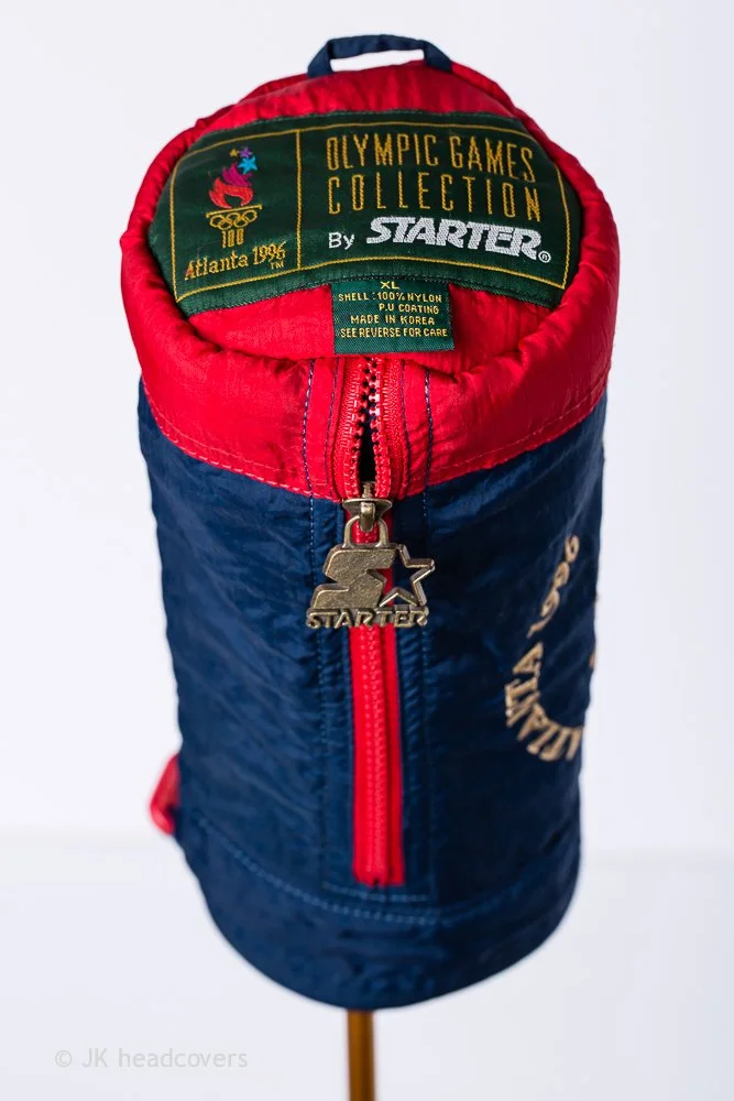A cylindrical bag resembling a sports jacket with red, blue, and green fabric. The top features an Olympic Games Collection label from 1996 Atlanta, with a zipper pull shaped like a gold star and athlete. The bag is designed for display or collection