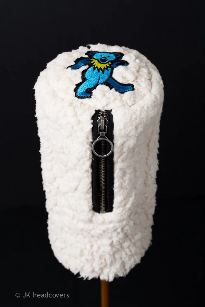 A white, fluffy golf headcover with a zipper in the front and an embroidered blue bear wearing a yellow collar on top, set against a black background.