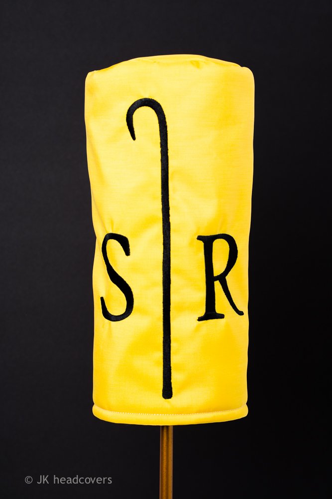 A yellow fabric headcover with the letters 'S' and 'R' and a black embroidered line in the middle, mounted on a wooden stick, against a black background.