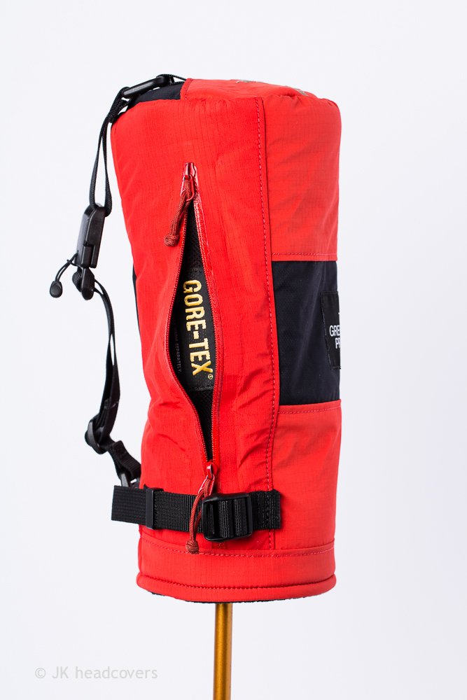 Red outdoor jacket with zipper pocket and black strap, labeled GORE-TEX, mounted on a stand.