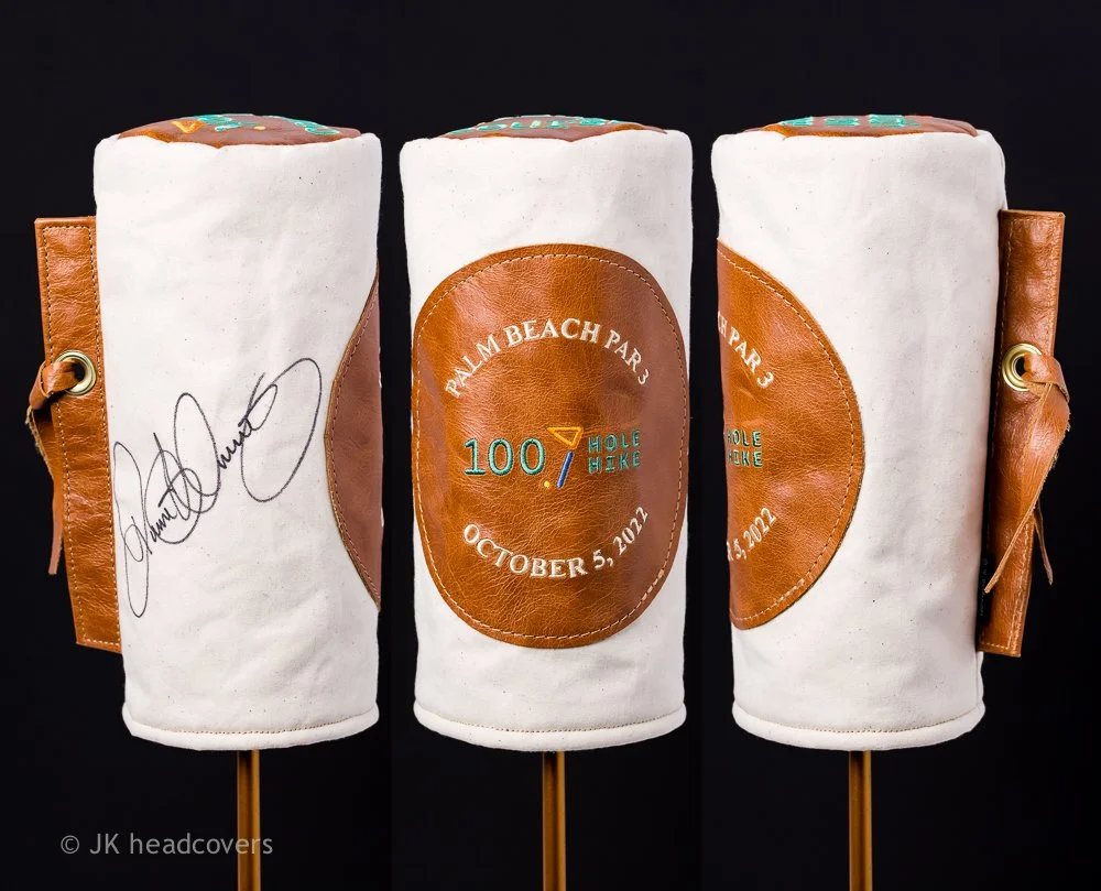 Three golf club head covers with a leather logo patch, one signed, against a black background. The middle cover has embroidered text for a golf event on October 5, 2022.