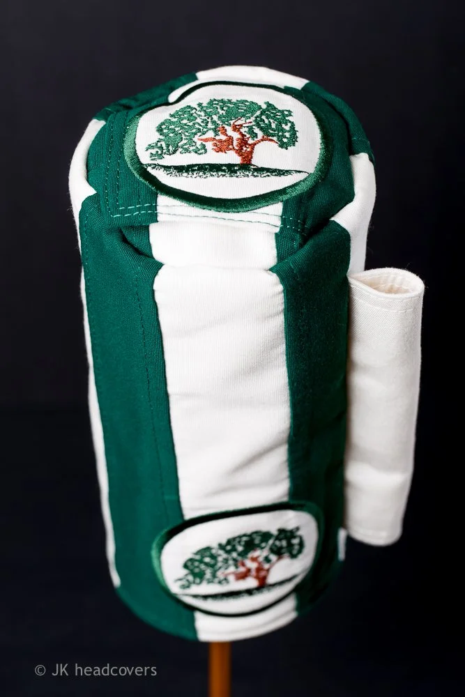 Golf club headcover with green and white stripes and a tree logo, standing vertically with a white towel rolled up beside it.