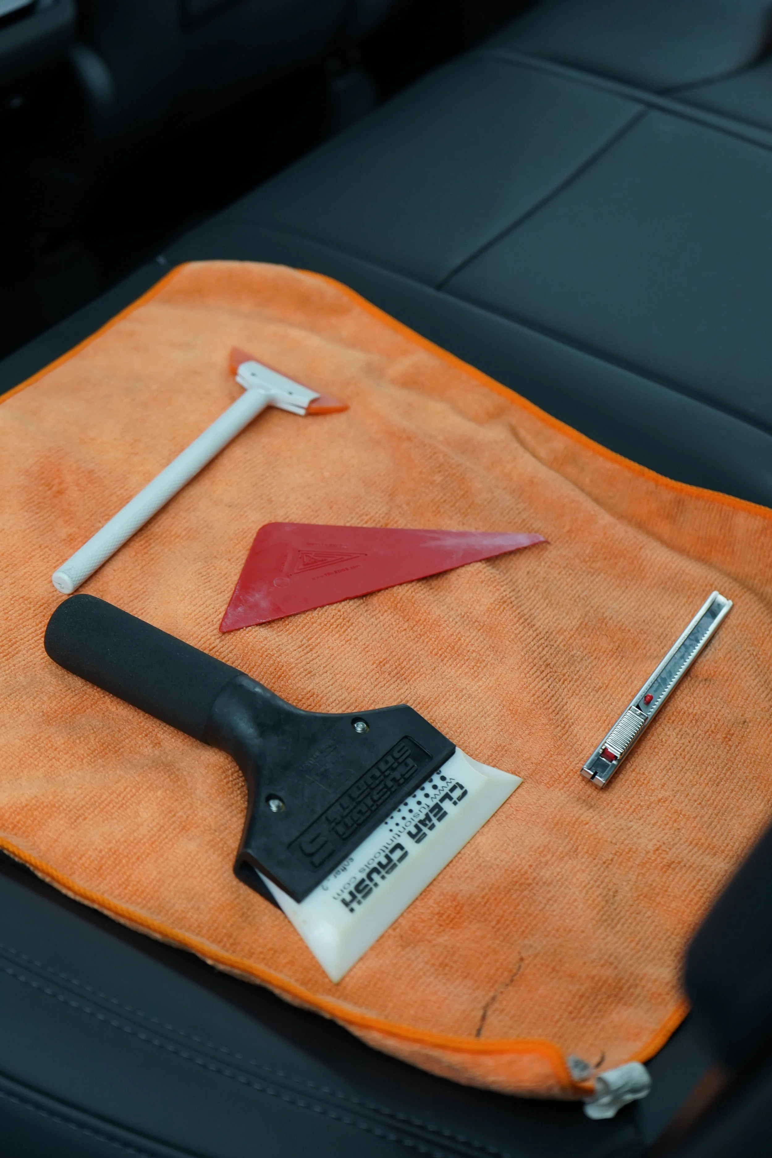 Car detailing tools on an orange towel, including a scraper, triangular plastic pry tool, razor knife, and a glue remover or cleaner.