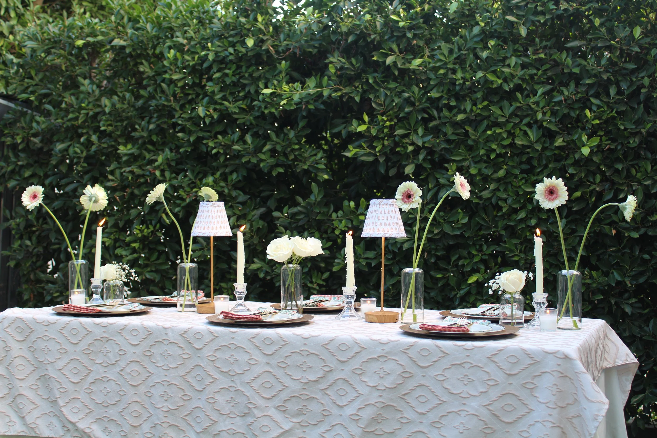 Outdoor dining table set with white tablecloth, floral centerpieces, tall candles, small lamps, and place settings, against a lush green hedge.