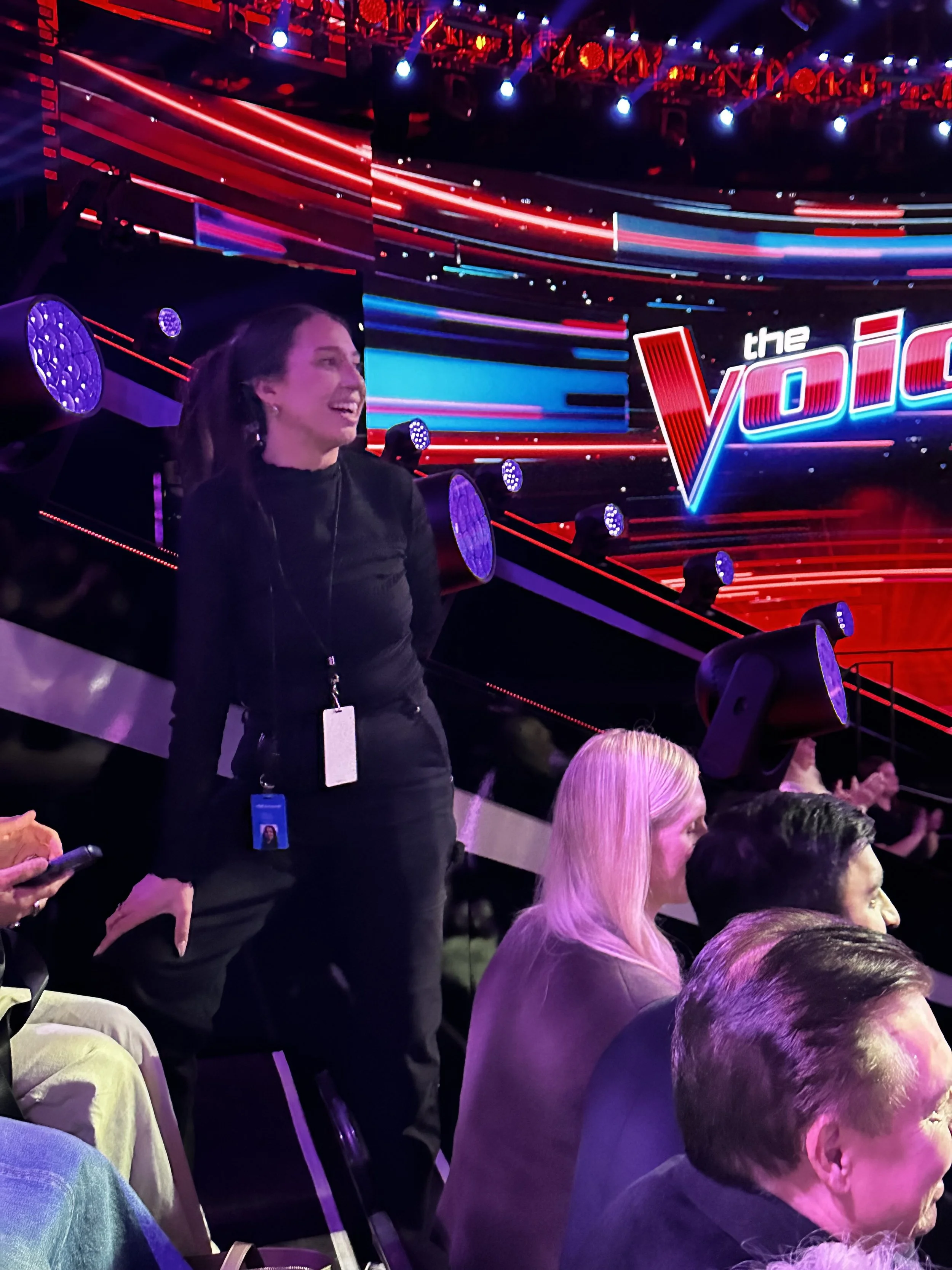A woman standing among seated people at a talent show stage with the logo 'The Voice' on a bright digital screen behind her.