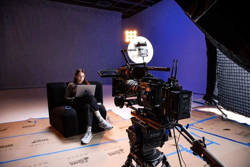 A woman sitting on a black armchair with a laptop in a studio setup, with professional film equipment and lighting around her.