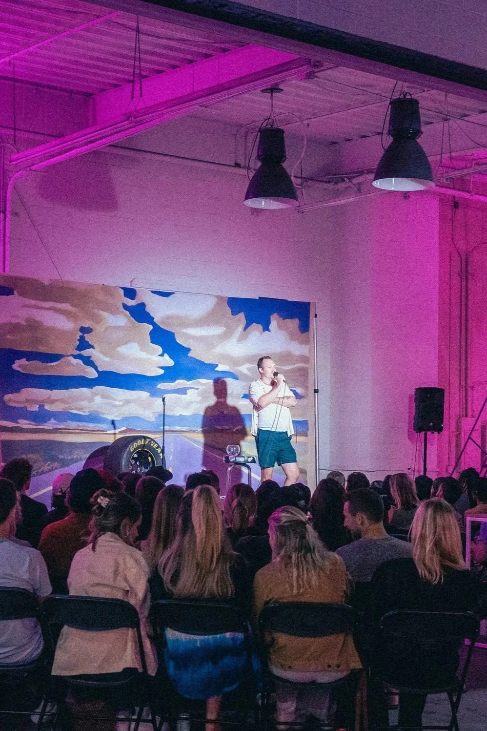 A man performs stand-up comedy on stage with a microphone, audience seated in front, a painted sky backdrop, and industrial lighting with pink hues.