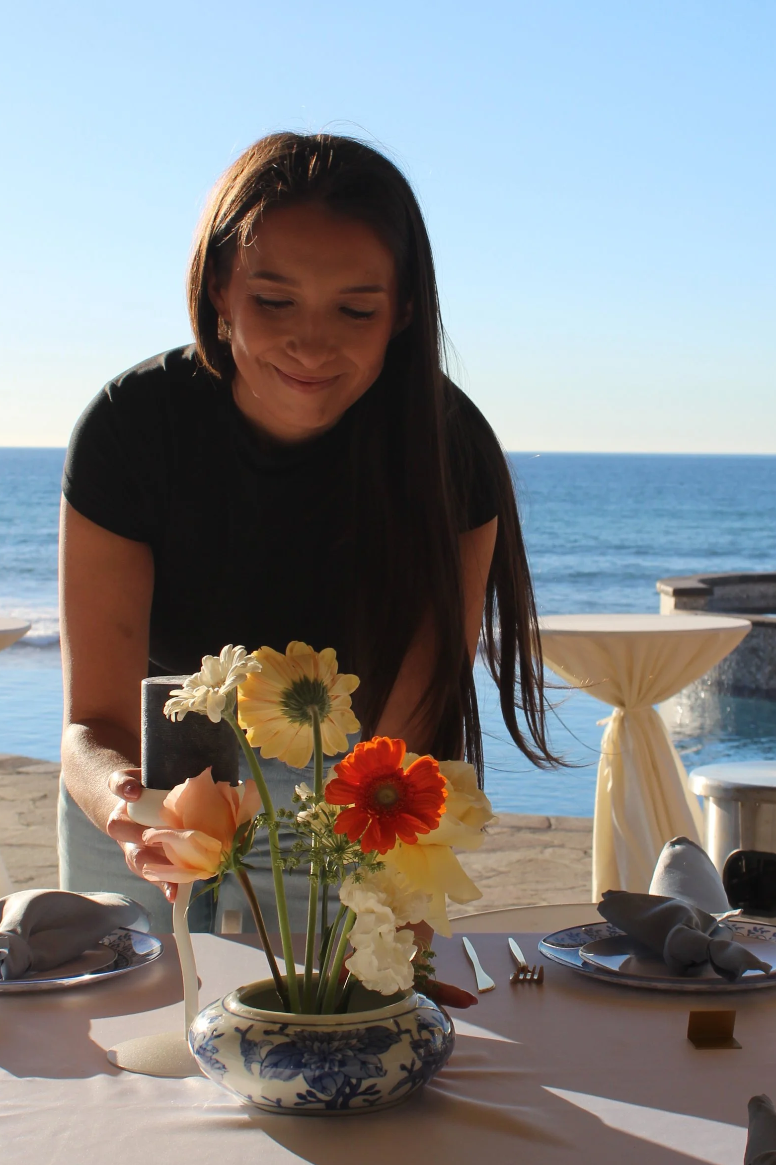 A woman in a black shirt arranging a vase of colorful flowers on a table near the ocean, with a clear blue sky in the background.