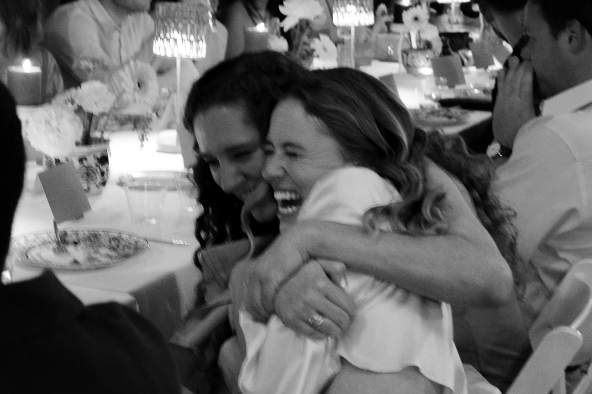 Two women sharing a laugh and hug while sitting at a table during a gathering or celebration.