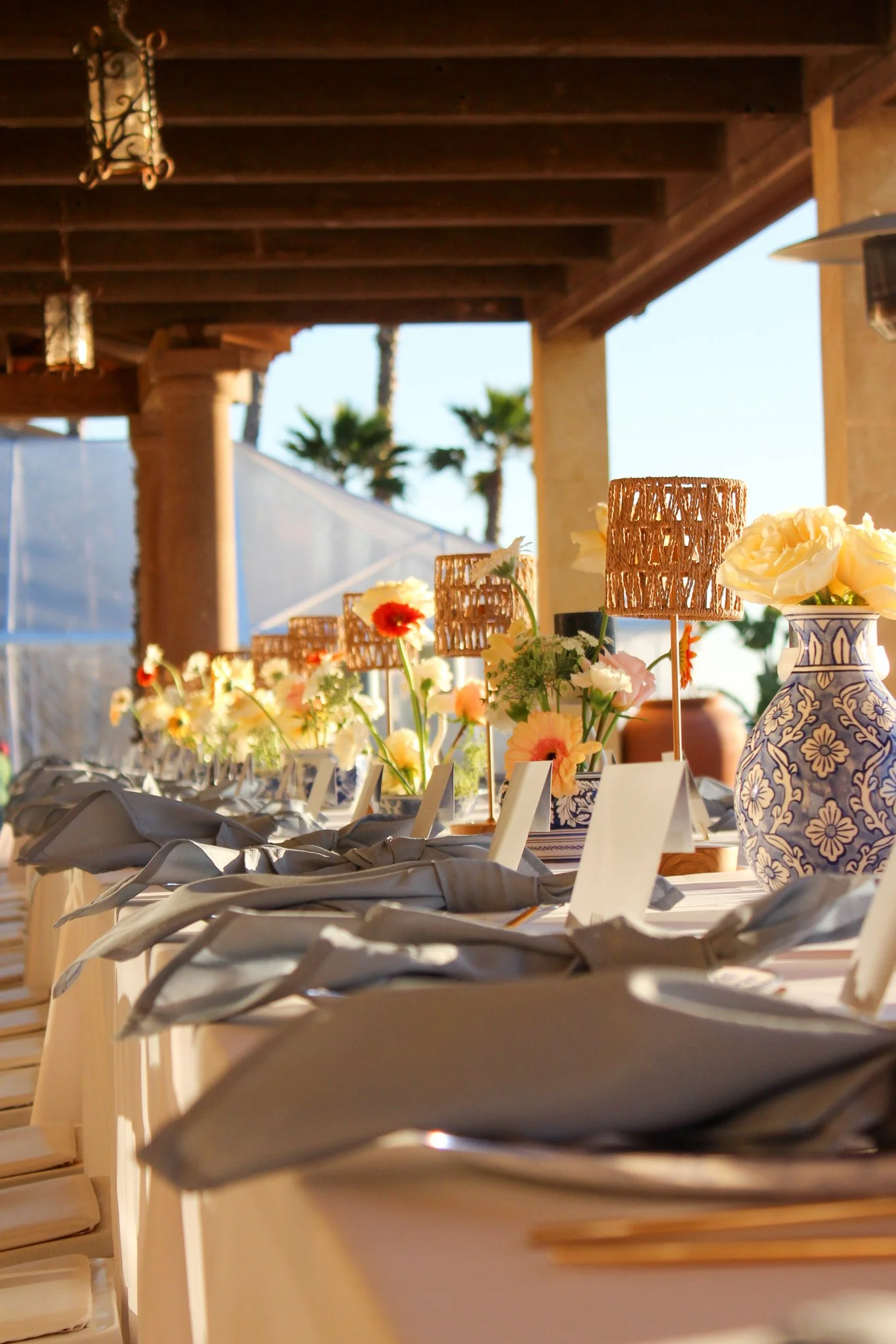 Long table set up outdoors with flower arrangements, woven lampshades, and place settings for a gathering or event, with a sunny sky and palm trees in the background.