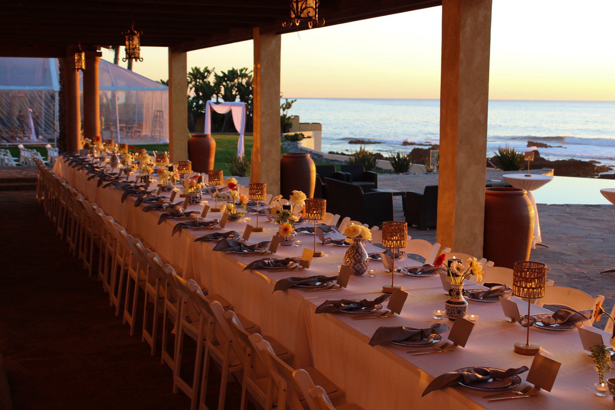 Long banquet table set for a dinner with flowers and candles, overlooking the beach at sunset with the ocean and sky in the background.